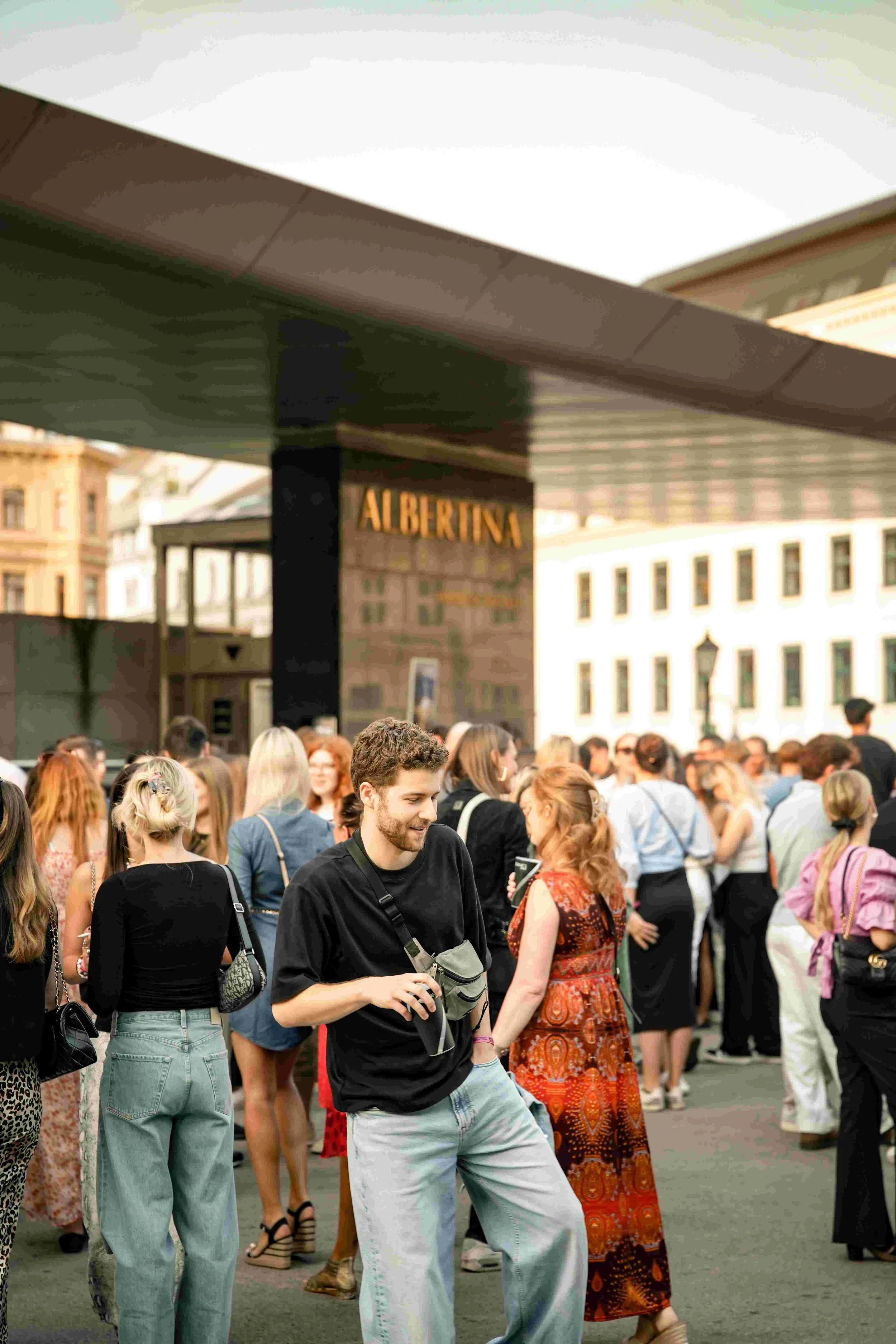 A crowd of people gathered outdoors in front of a building with a sign that reads 'ALBERTINA.' The individuals are chatting and appear to be at an event or gathering, dressed casually with some using smartphones.