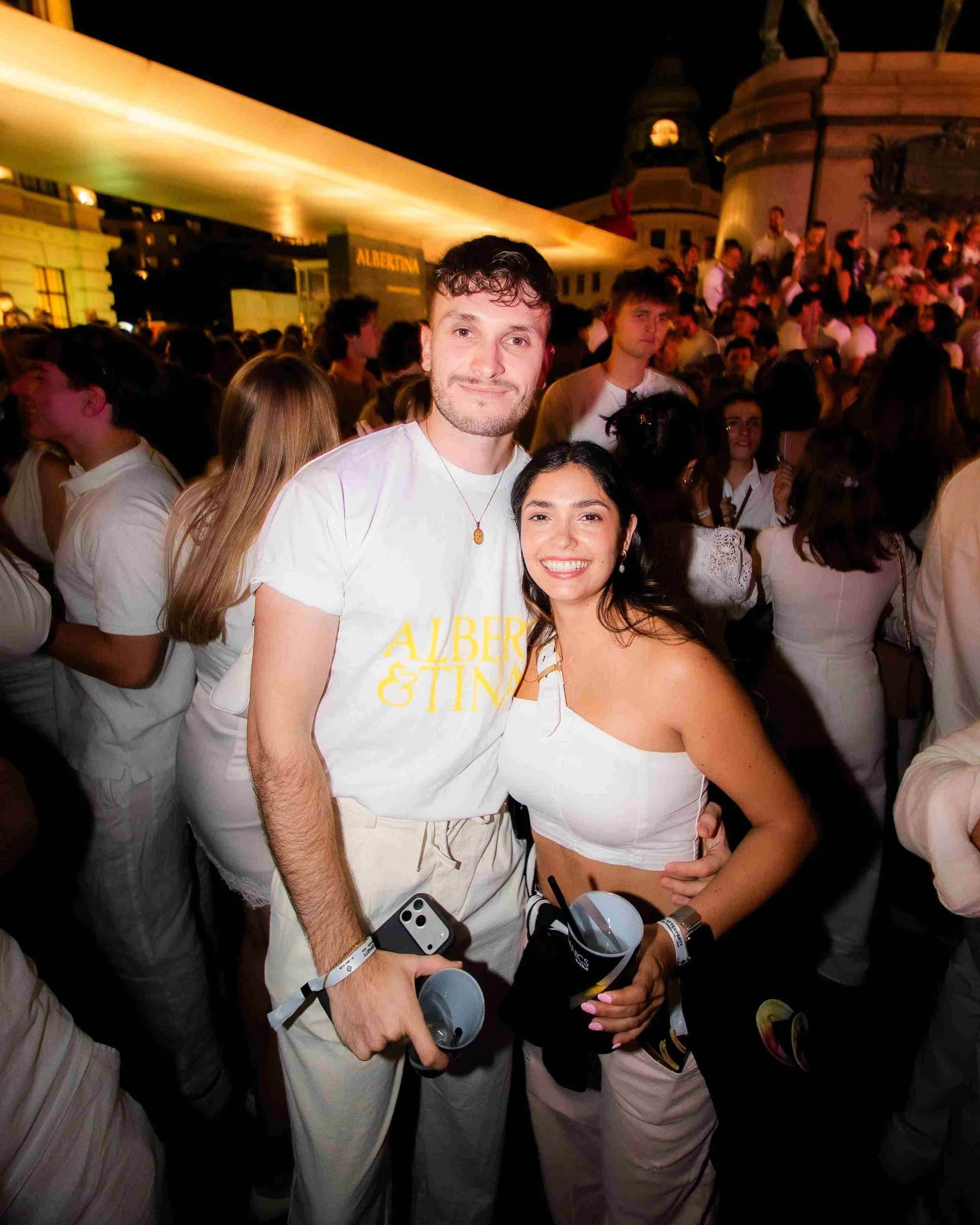 Two smiling people, a man and a woman, pose in a crowded, nighttime outdoor event, with many people around them and a building with a rounded rooftop in the background.