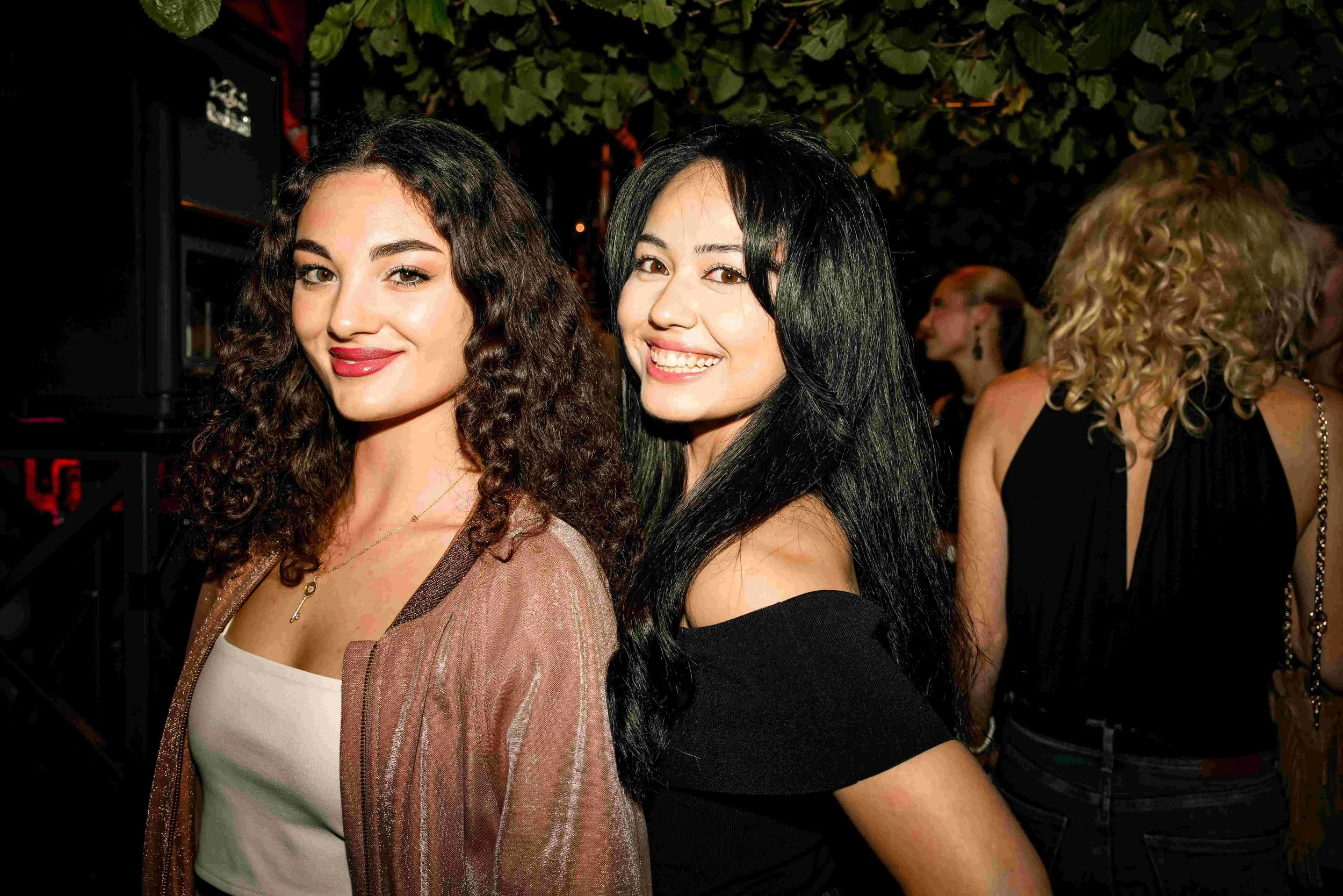 Two smiling women with curly and straight black hair, wearing stylish outfits, at a social event at night with greenery and other guests in the background.