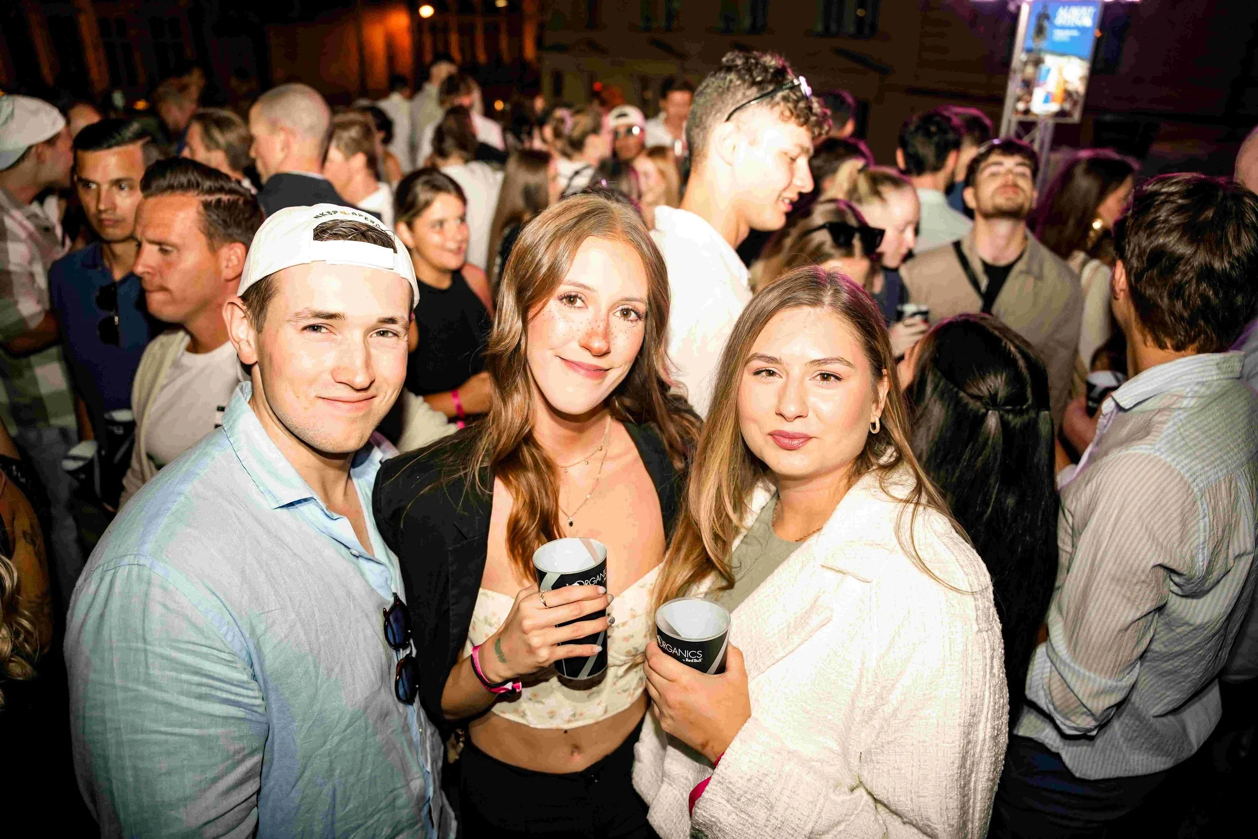 Group of young people at a crowded indoor event, some holding cups, smiling and engaging with each other.