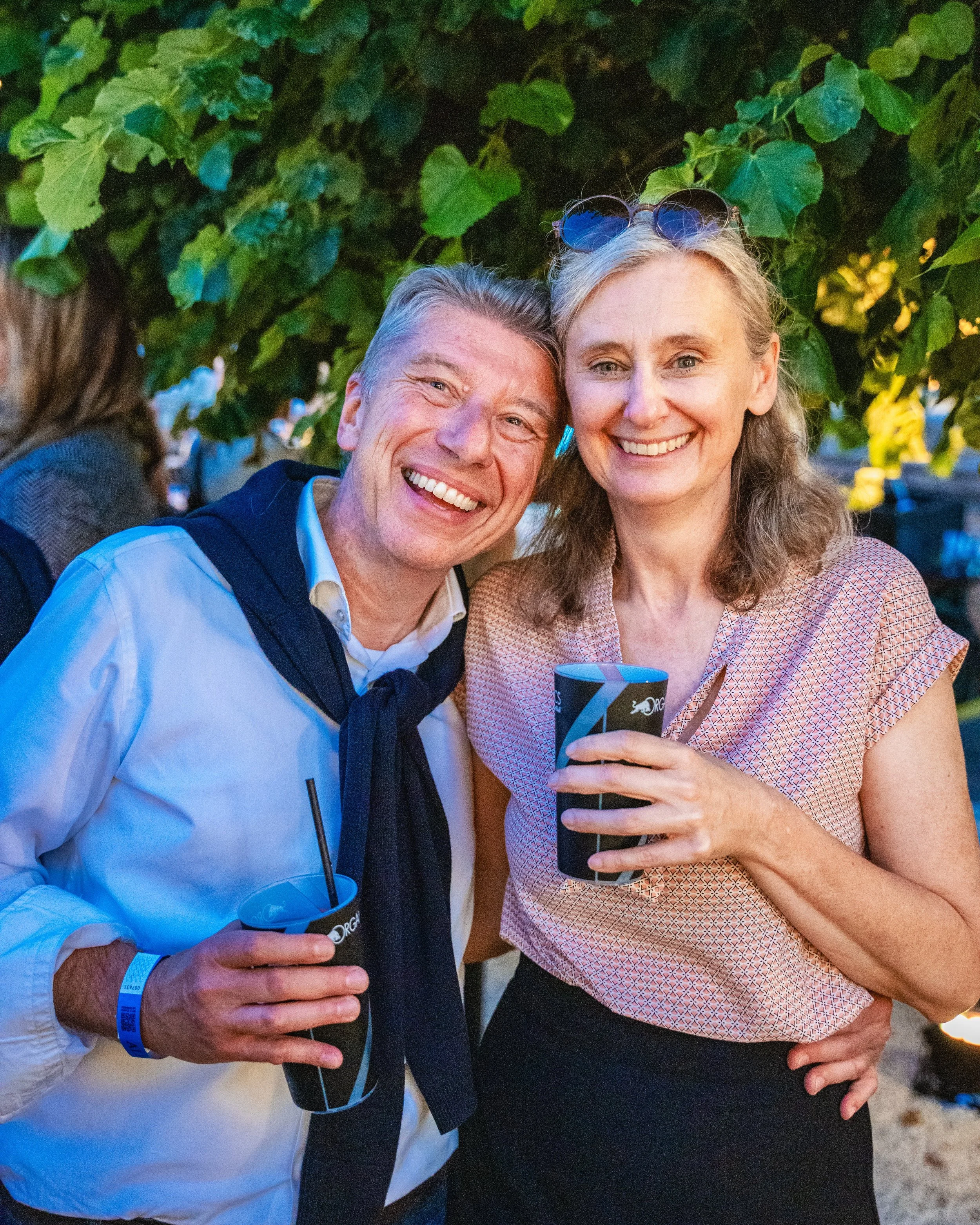 Two smiling women at an outdoor gathering, holding drinks and standing close to each other under a leafy tree. One woman is wearing sunglasses on her head and a light-colored patterned blouse, while the other is dressed in a white shirt with a dark v