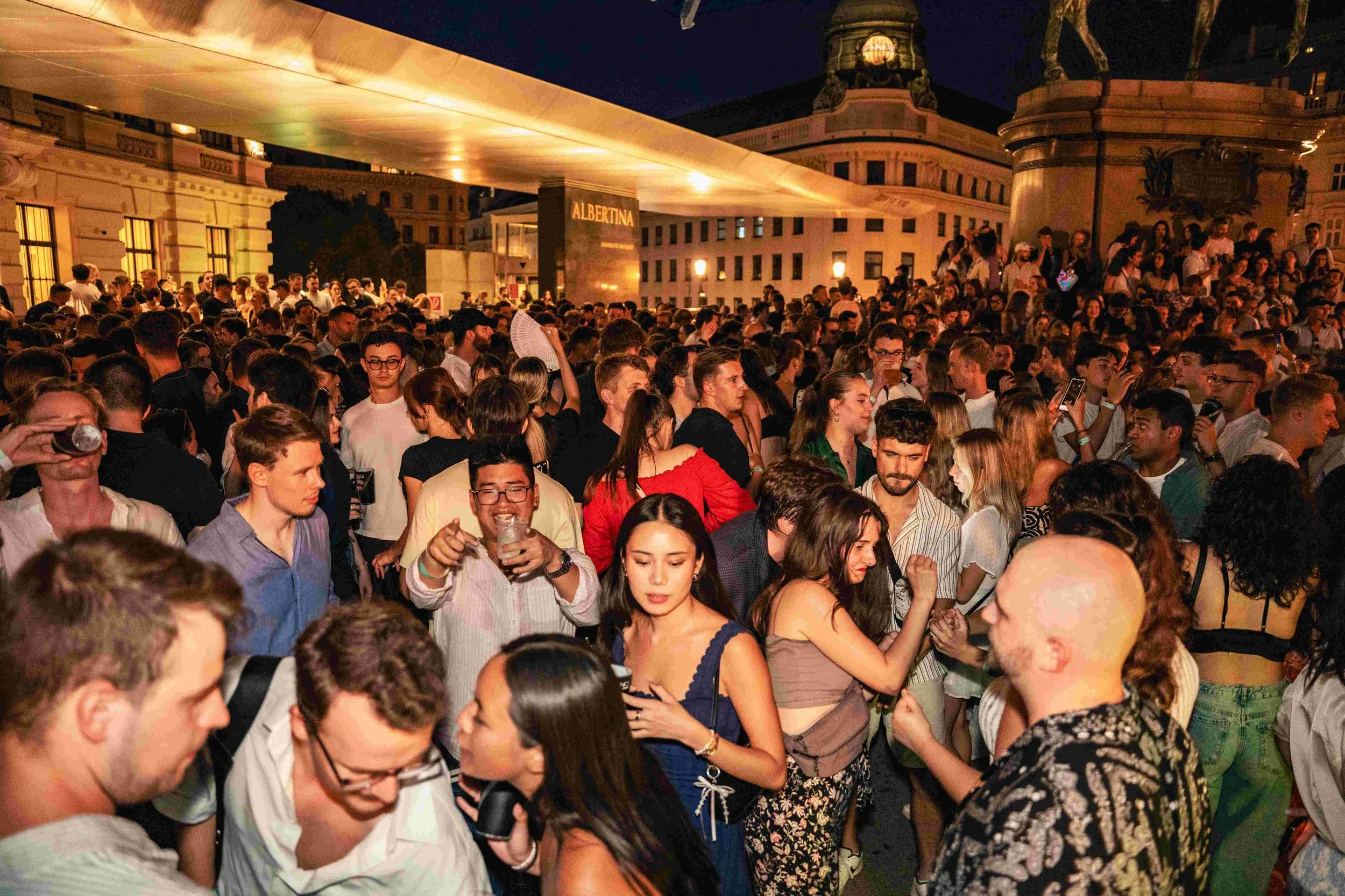 A large crowd of people gathered outdoors at night, socializing and enjoying the event. The background features the building with the illuminated sign reading 'ALBERTINA' and some architectural details.