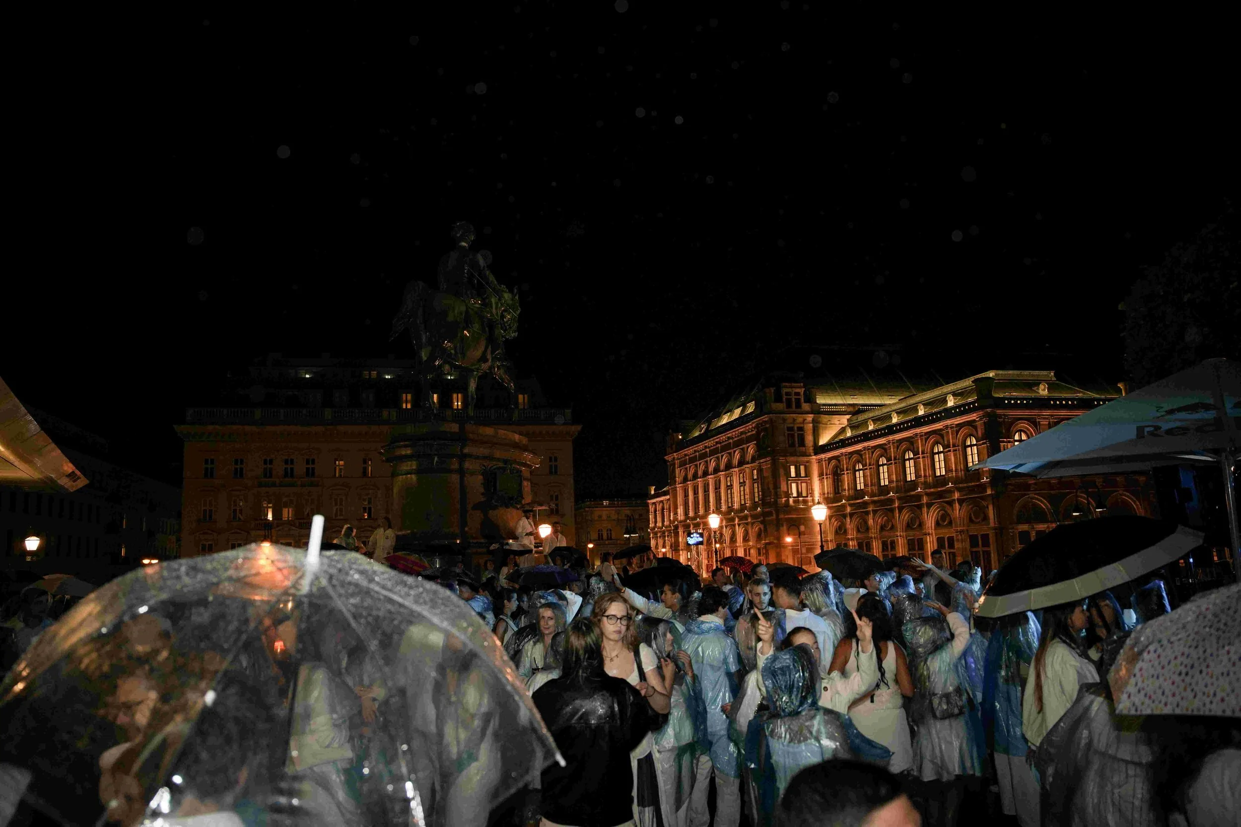 A crowd of people gathered in a city square during a rainy night, many holding umbrellas. In the background, historic buildings and a statue of a person on horseback are illuminated, with a dark, starry sky above.