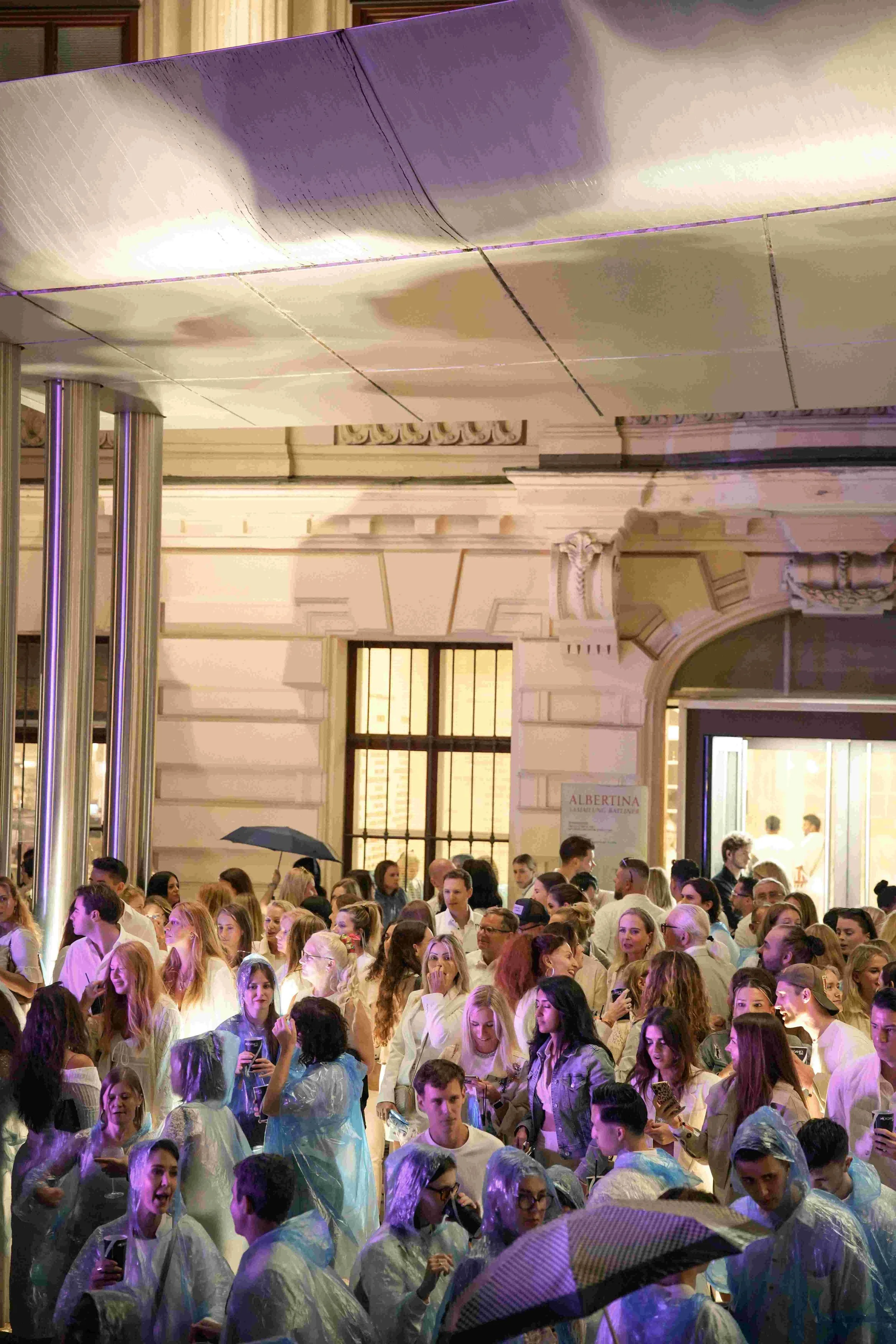 Crowd of people gathered indoors, many wearing rain ponchos, with some umbrellas visible, under soft lighting and a decorated interior with arches and columns, possibly at an event or celebration.