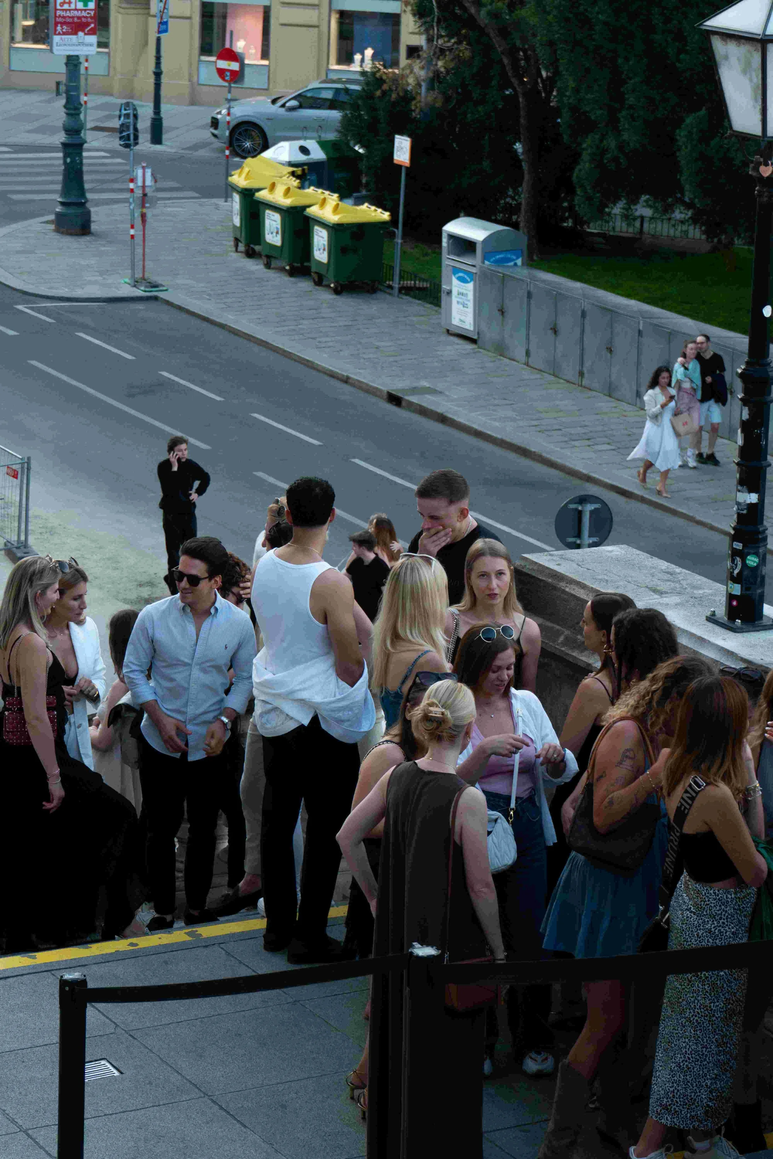 People waiting in line outdoors on a city sidewalk, with some talking and others looking at their phones. There are trash bins, street signs, and a parked car in the background.