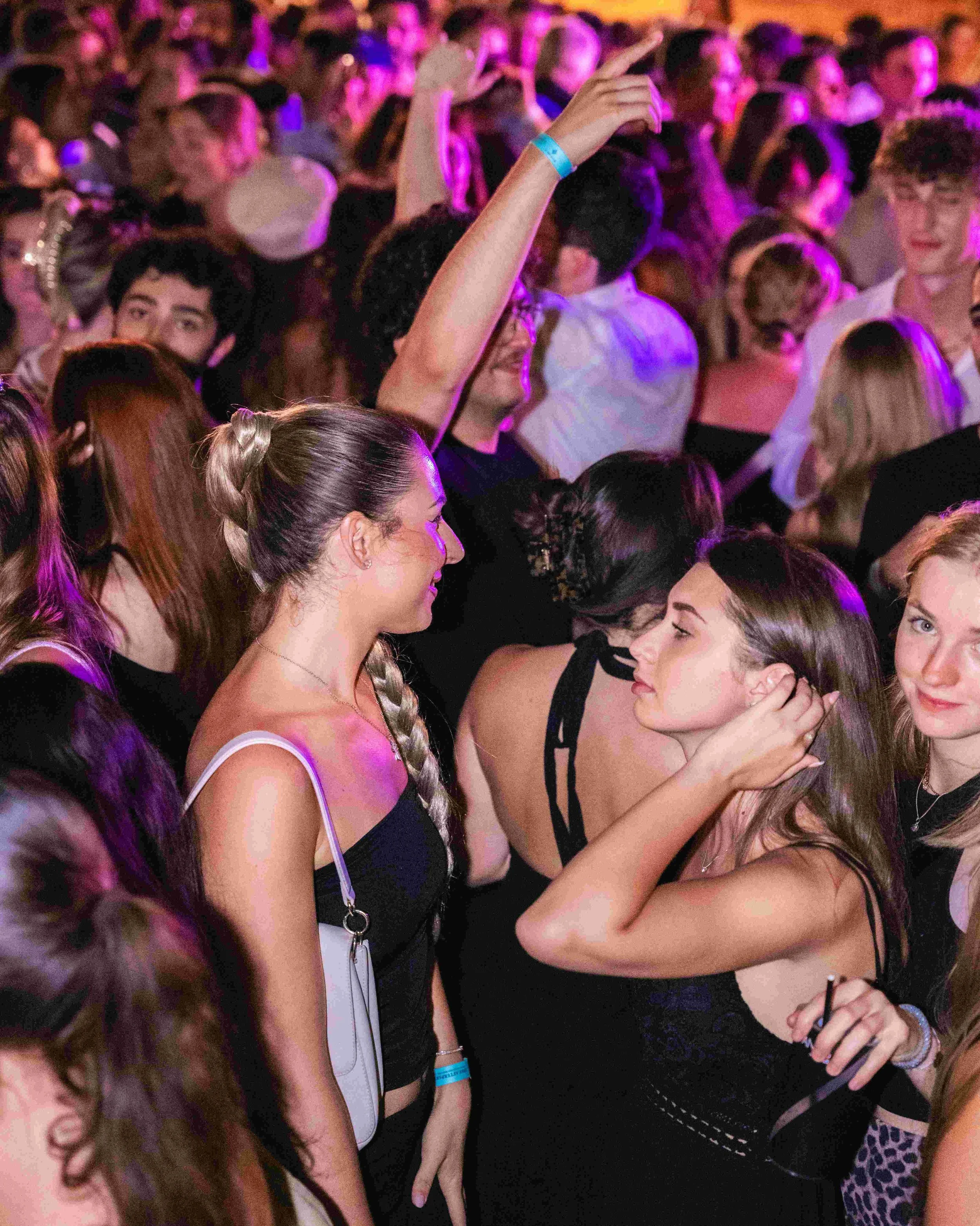 A crowded dance floor with people socializing, some women hugging, others dancing or talking, with colorful lighting creating a vibrant atmosphere.