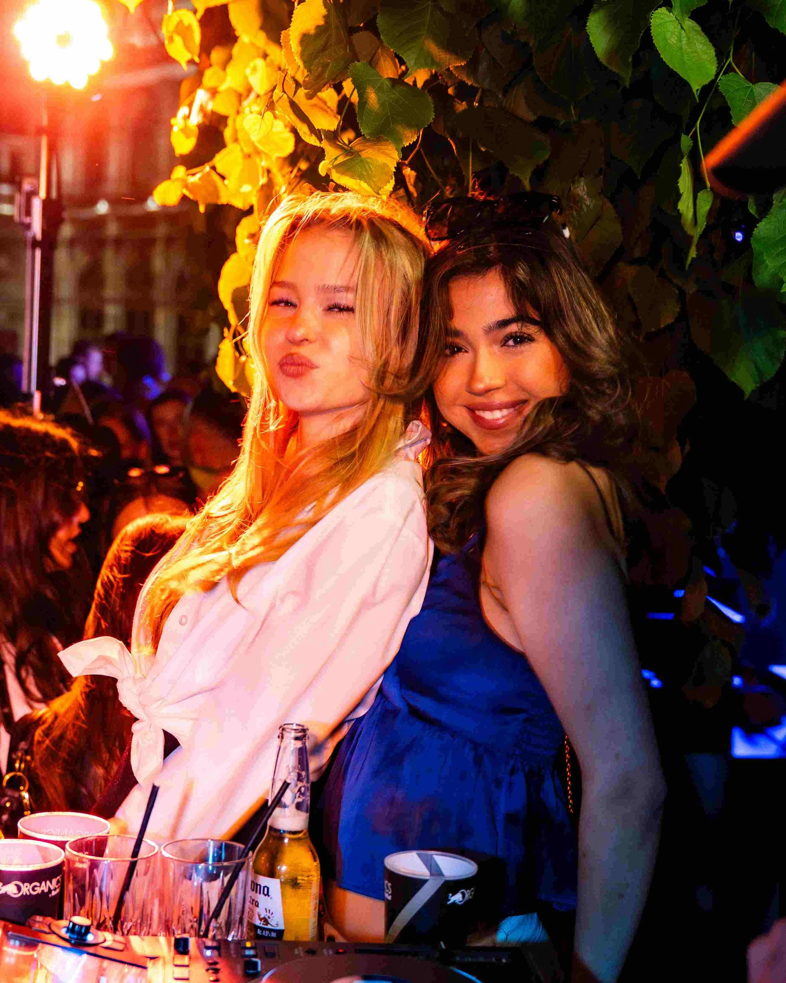 Two young women hugging and smiling at a lively party with colorful lighting and drinks on the table.