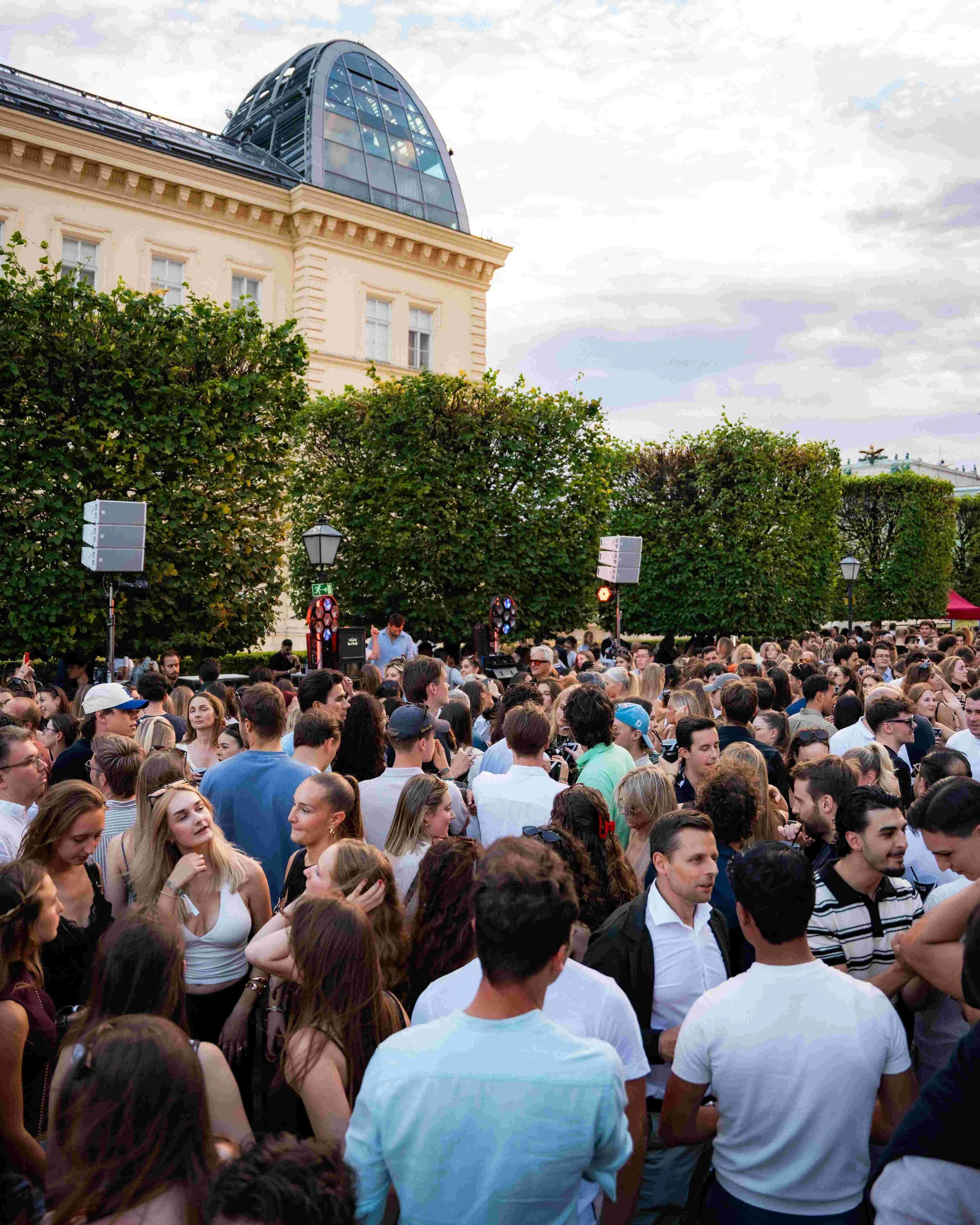A large crowd of people gathered outdoors in an urban area, with some people talking and looking around. In the background, there are green trees, a beige building with classical architecture, and cloudy sky above.