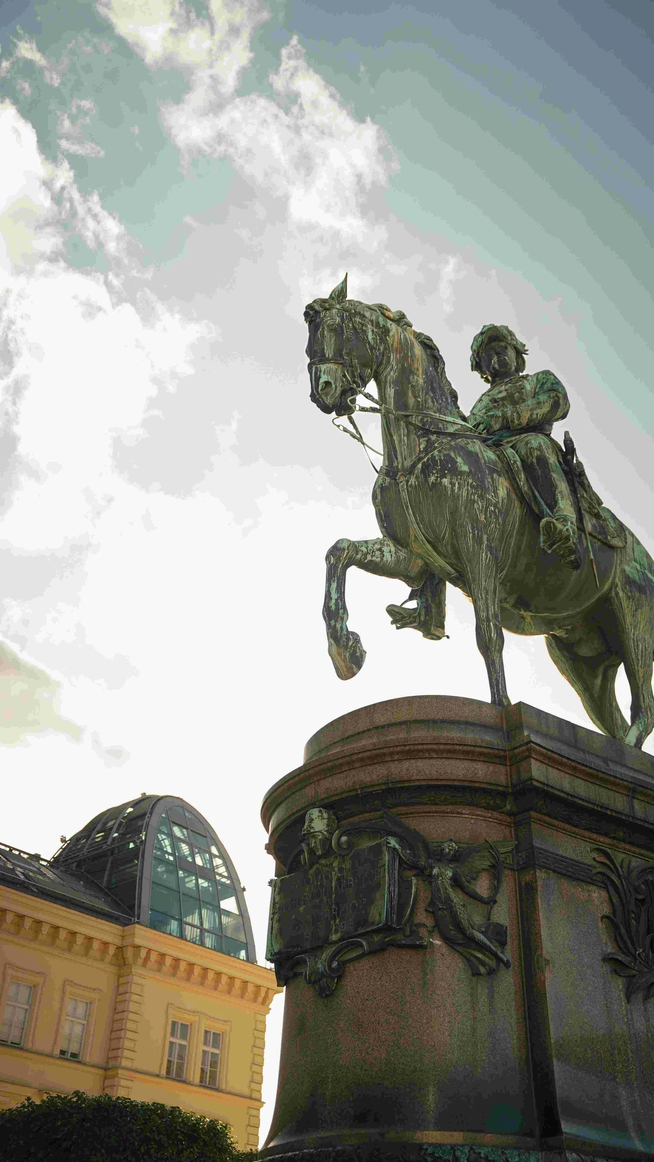 A bronze equestrian statue of a historical figure on horseback, located outdoors against a cloudy sky, with a building and a glass-domed structure nearby.