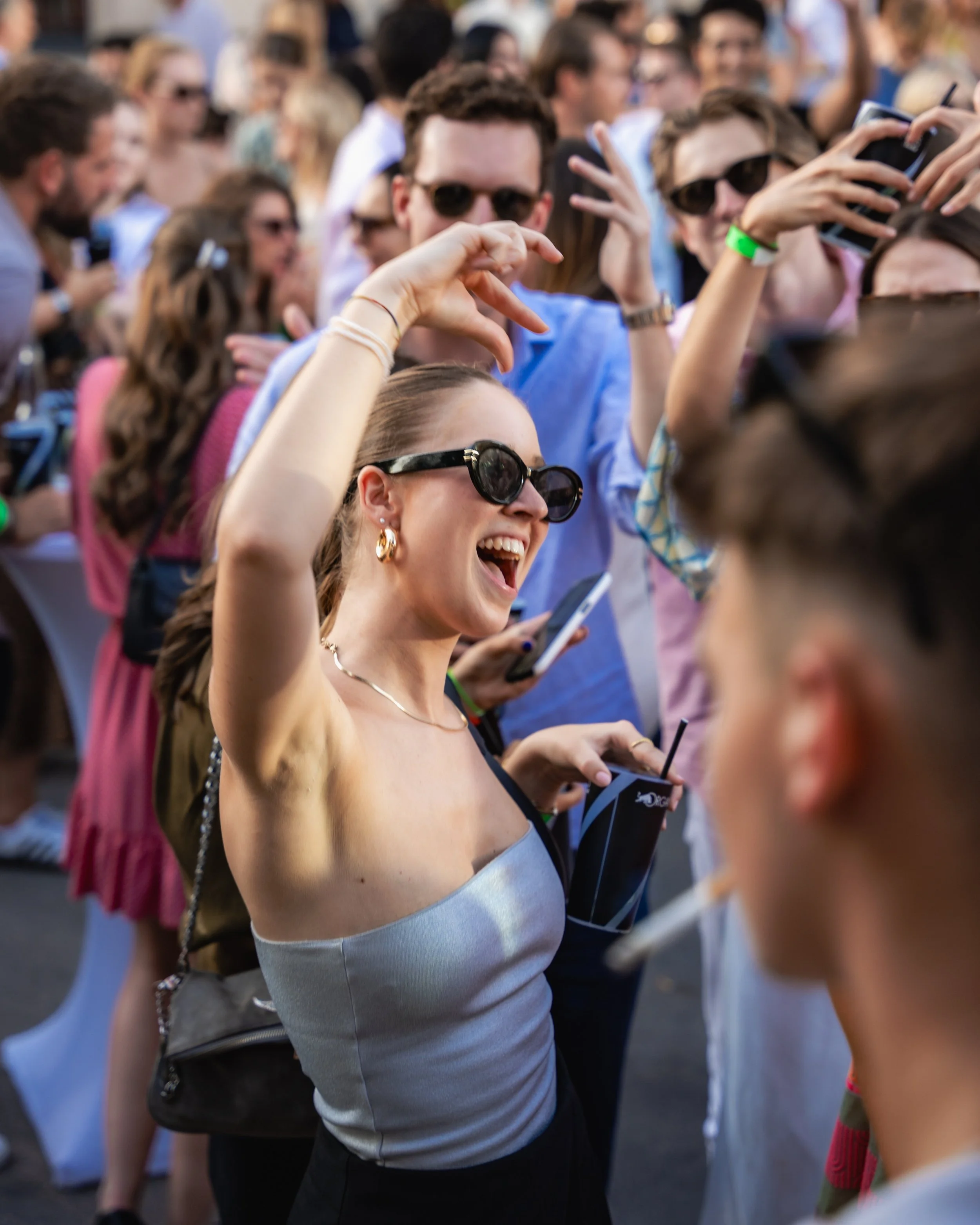 A group of young adults at an outdoor event, many wearing sunglasses, standing close together and smiling while using their smartphones.