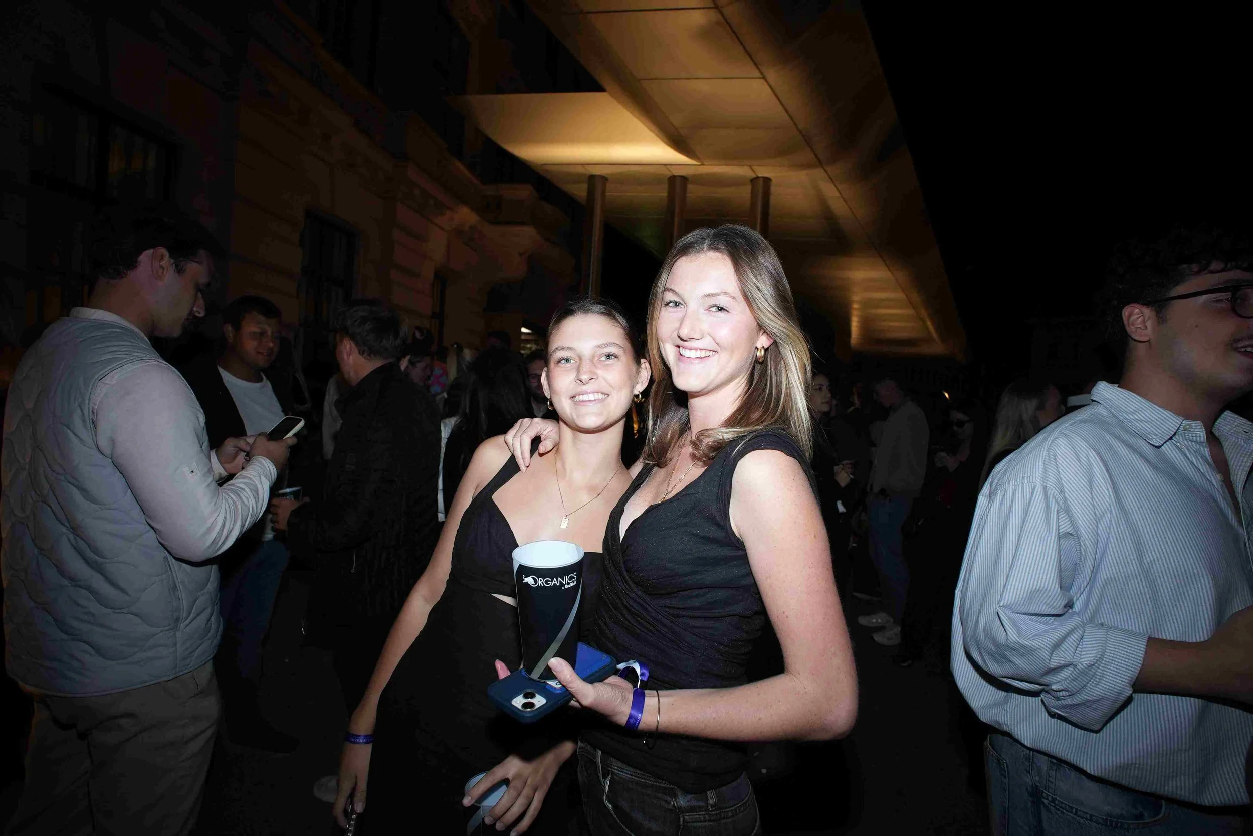 Two women smiling at the camera at a social event, with other people in the background, holding drinks, inside a dimly lit venue with wooden interior.
