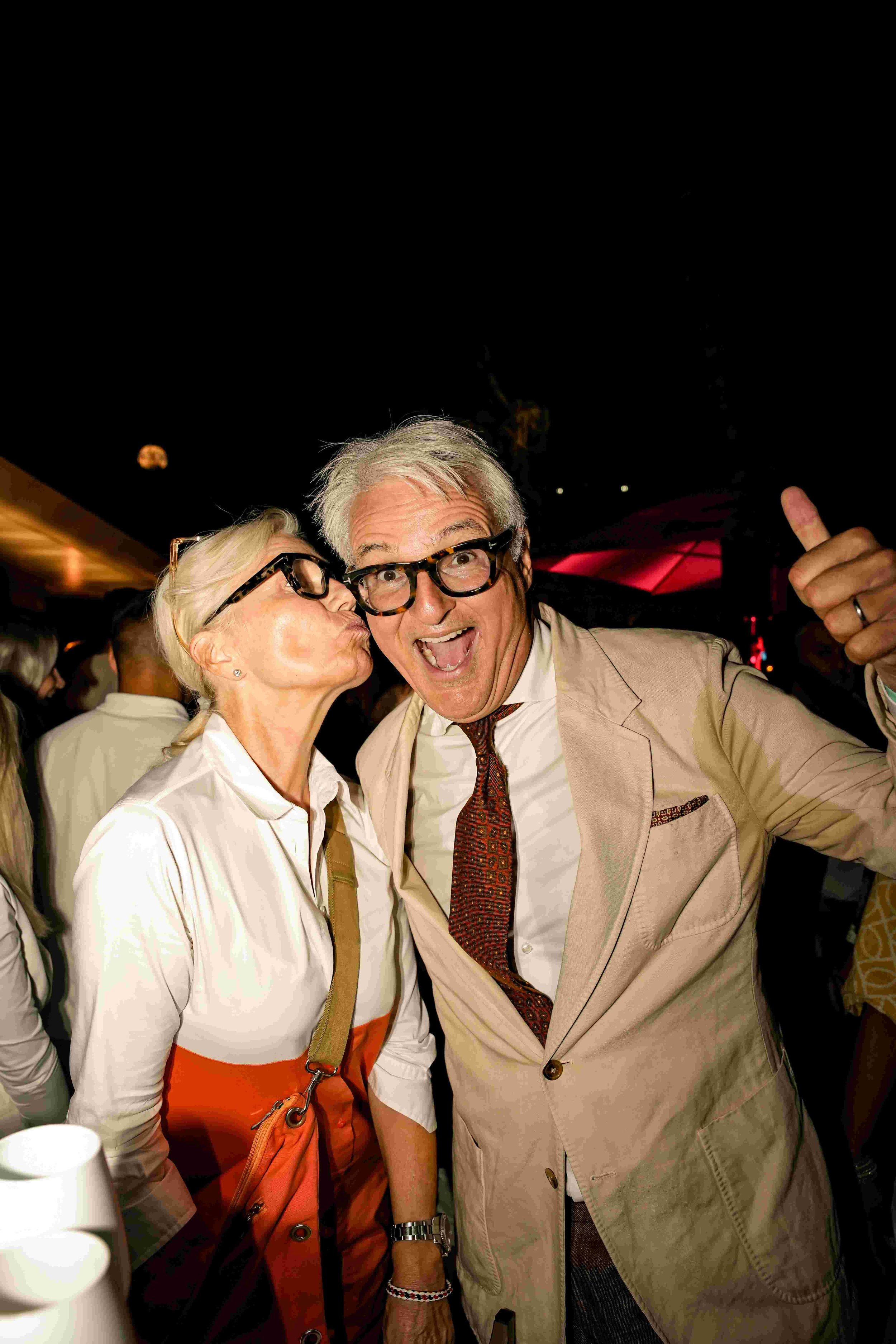 A woman with short blonde hair and glasses kisses a man with gray hair and glasses on the cheek at a social event. The man is smiling and giving a thumbs-up gesture.