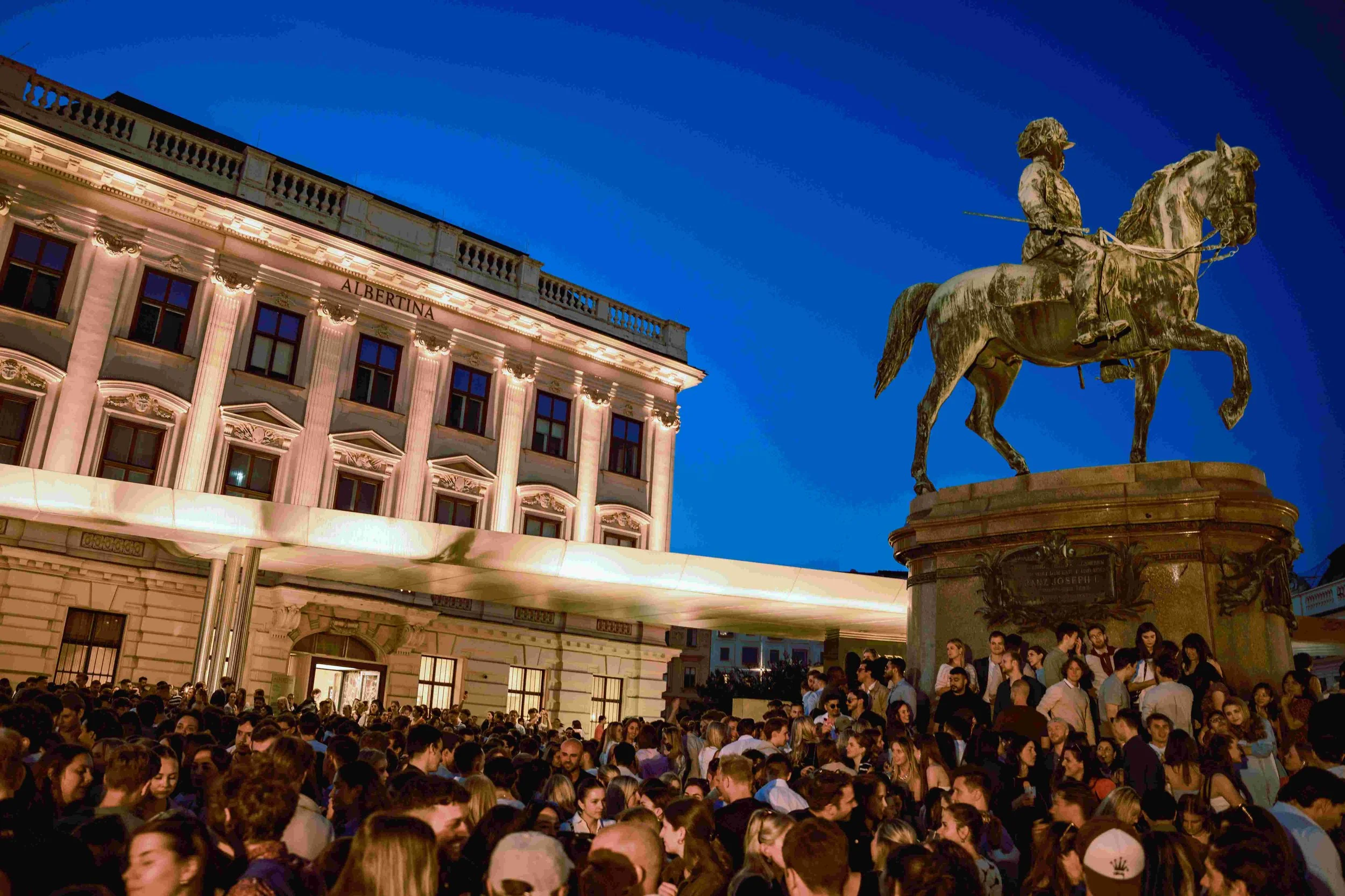 Crowd gathered in a city square at night with a historic building and an equestrian statue visible in the background.