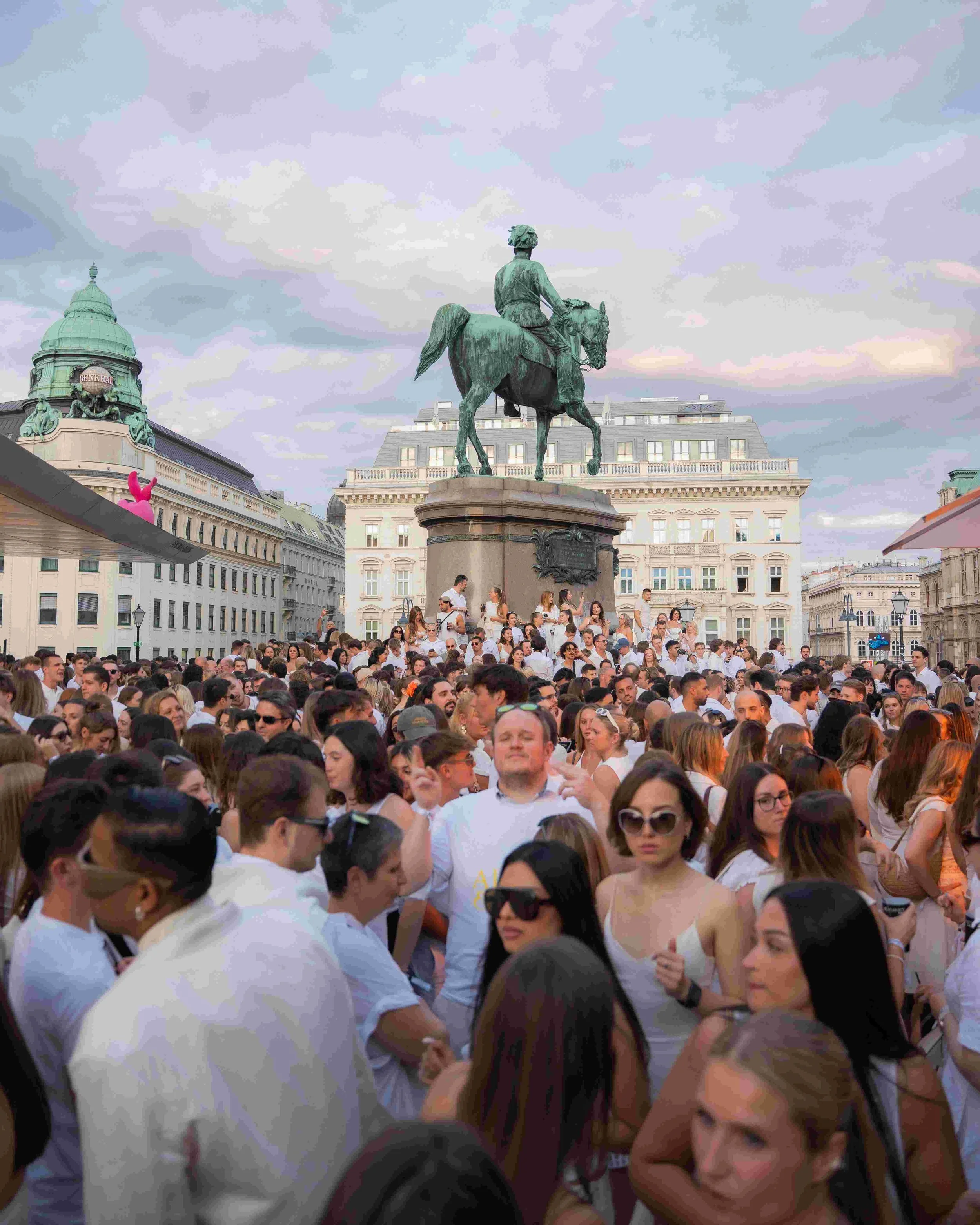 Crowd of people gathered around an equestrian statue in an urban square with historic buildings and cloudy sky in the background.