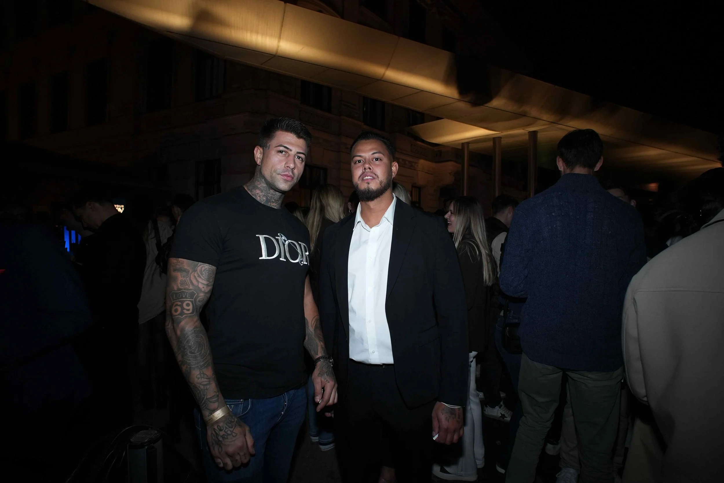 Two men standing together at a nighttime event, with a crowd in the background. One man has tattoos on his arms and neck, wearing a black Dior t-shirt, and the other is dressed in a black blazer and white shirt.
