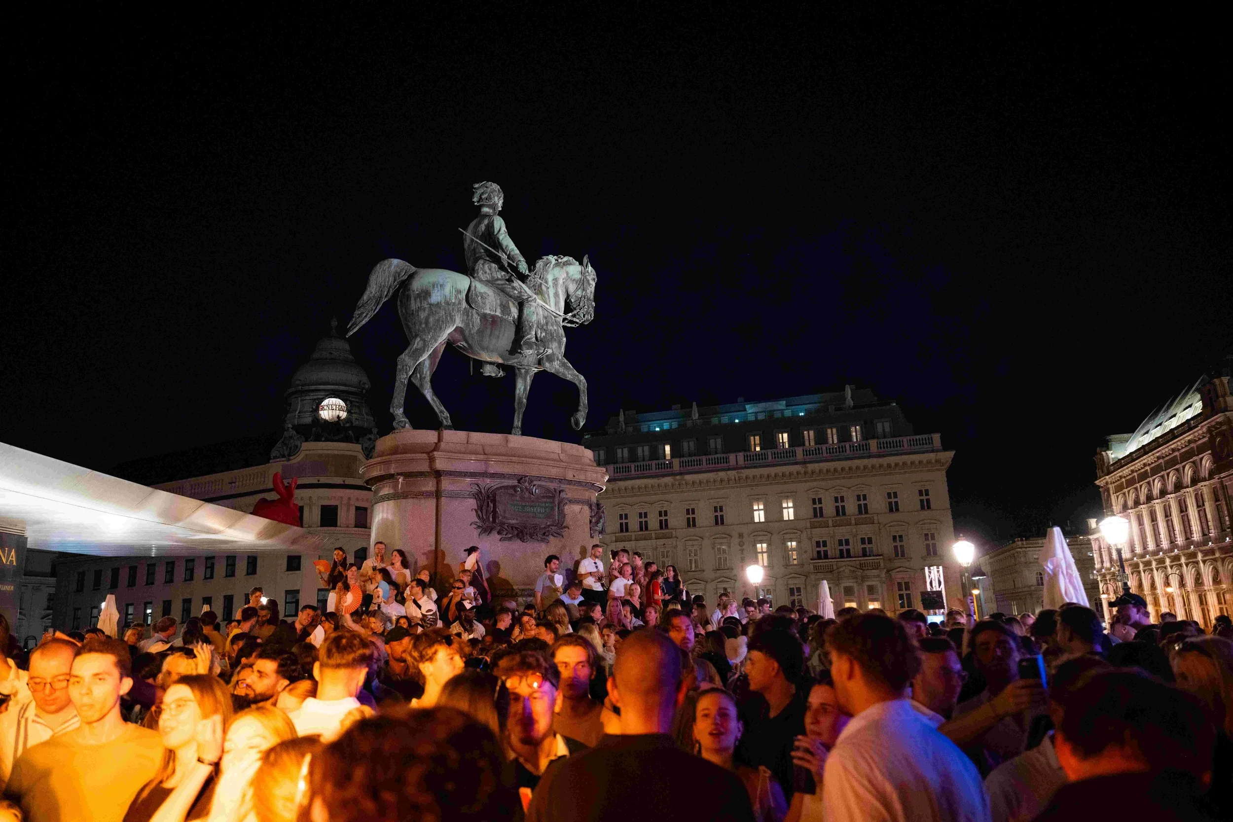 Nighttime scene at a crowded square with a large equestrian statue of a historical figure on horseback, illuminated against the dark sky. Surrounding buildings are lit, and there are many people gathered, some taking photos and enjoying the event.