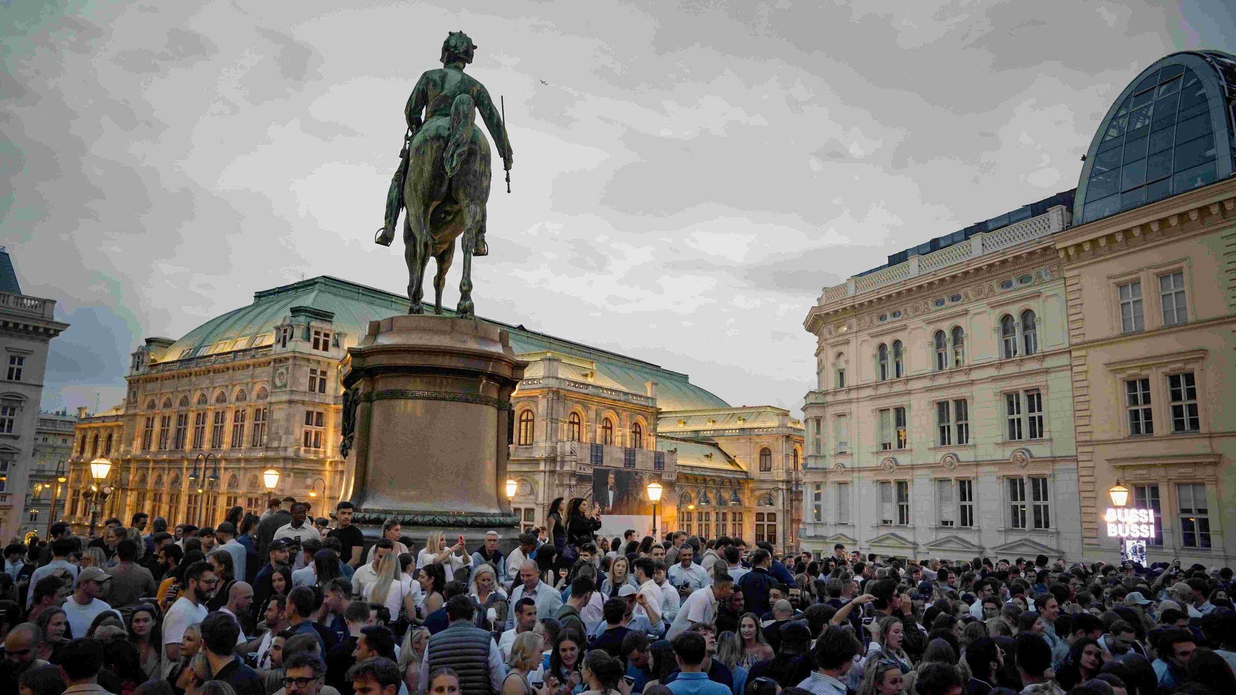 Crowds gathered in a city square with historic architecture and a bronze equestrian statue of a man on horseback in the center.