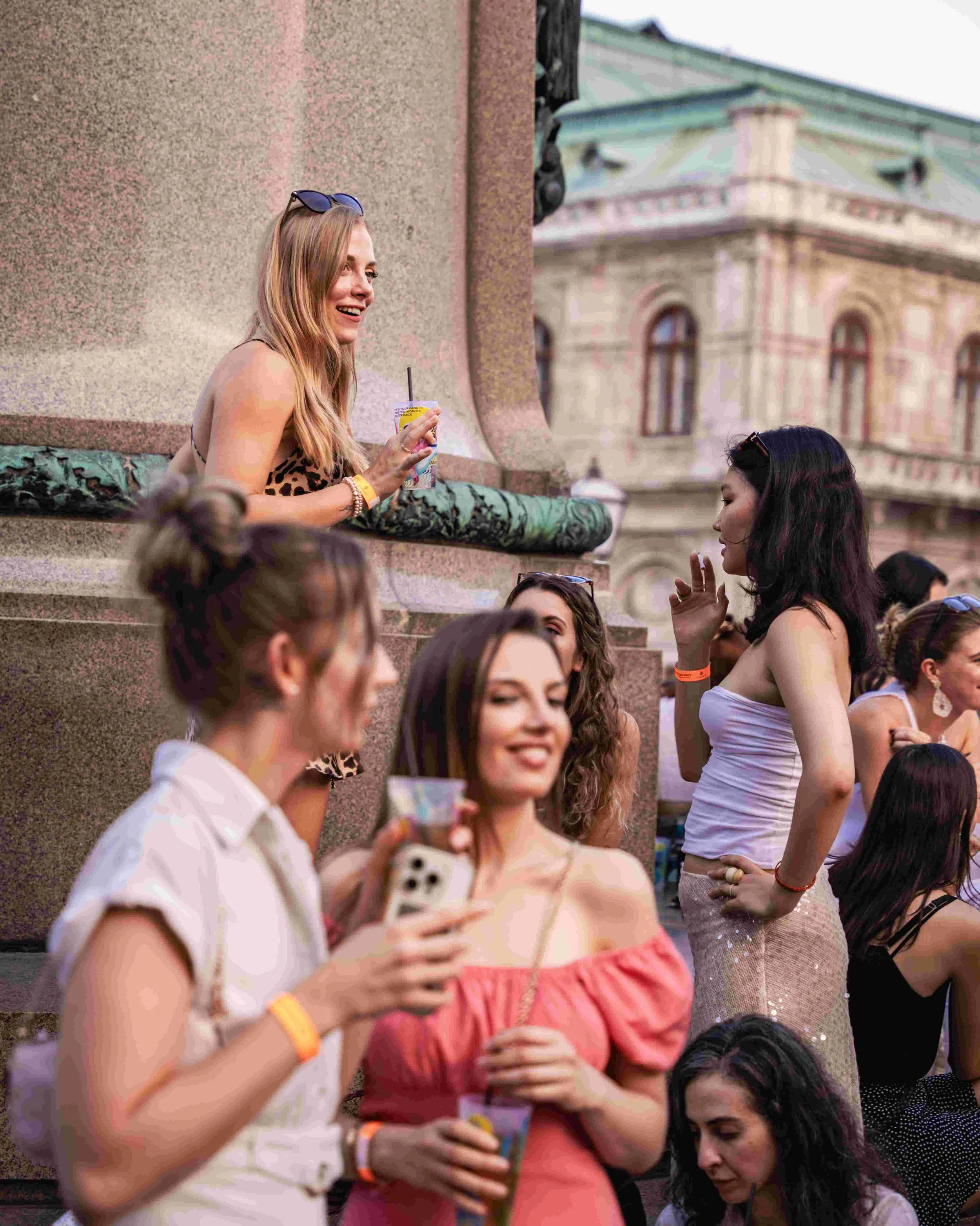 Group of young women socializing outdoors near historic building, some holding drinks, talking, and smiling.