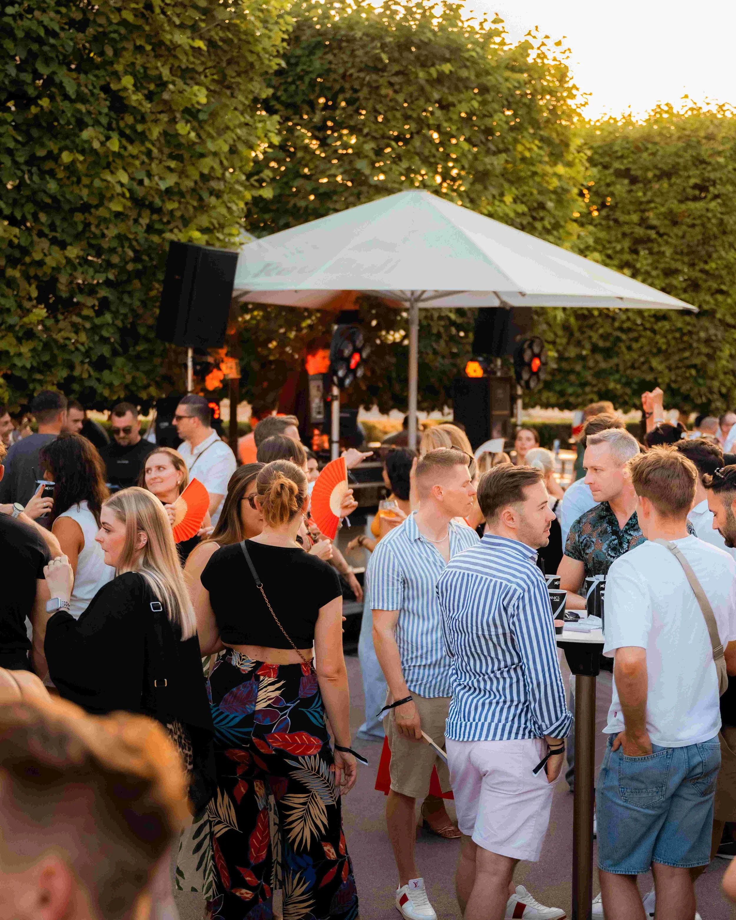 Crowd of people gathered outdoors at an event with a white canopy tent and green leafy trees in the background during sunset.