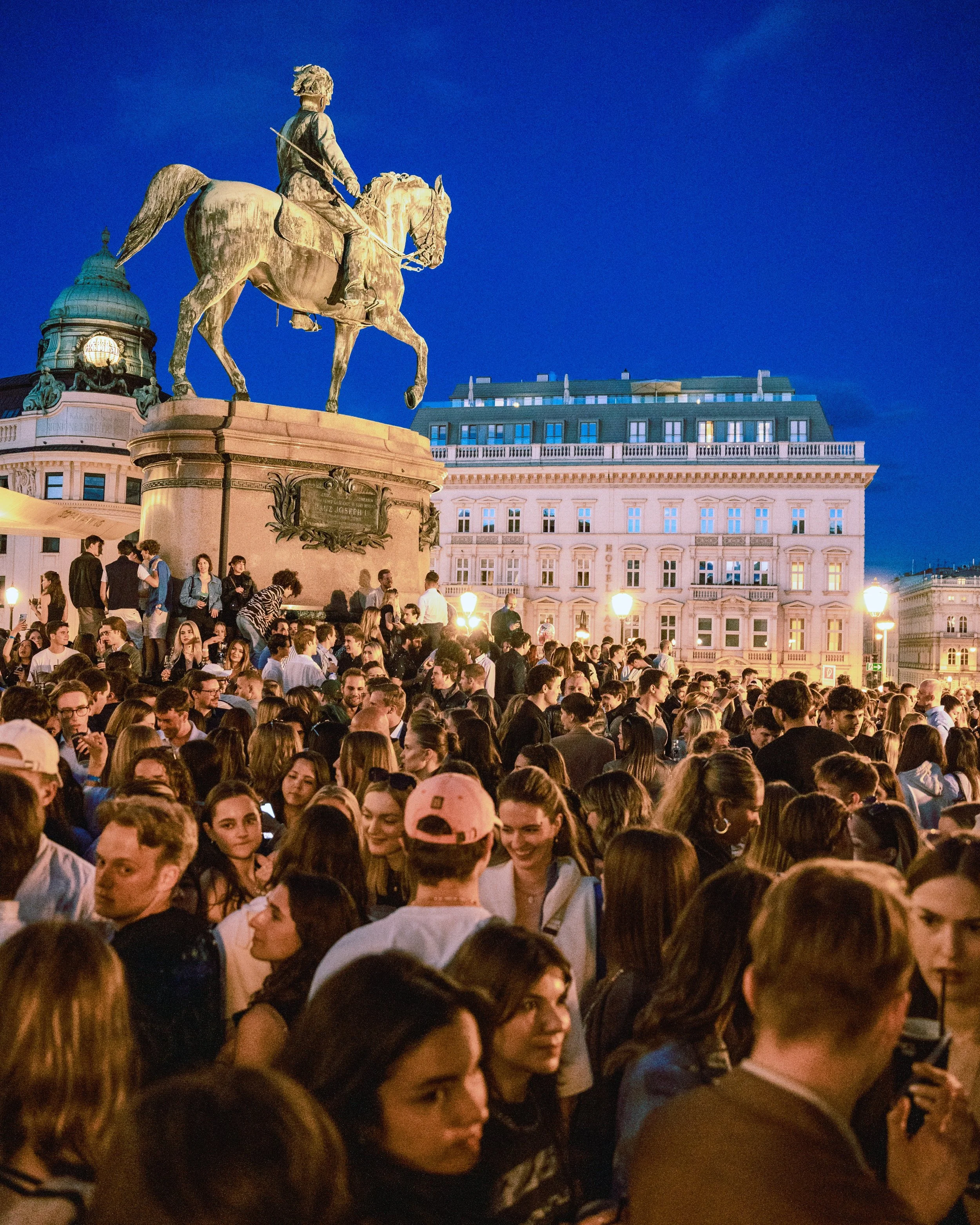 A large crowd of people gathered outdoors at night beneath a large statue of a man on horseback, with a historic building with multiple windows and a dome in the background.