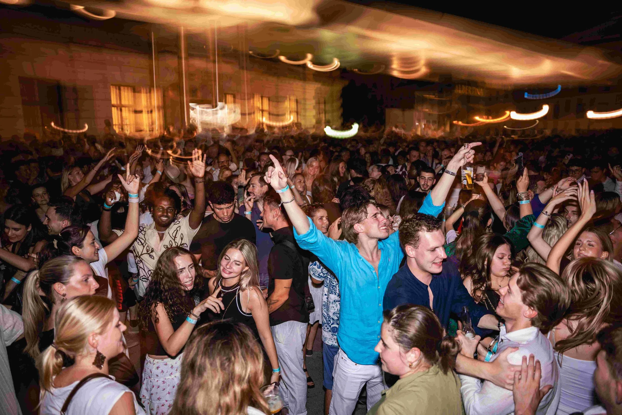 A crowded dance floor at a concert or party with many people smiling, dancing, and enjoying themselves. Colorful lighting creates swirling patterns on the ceiling.