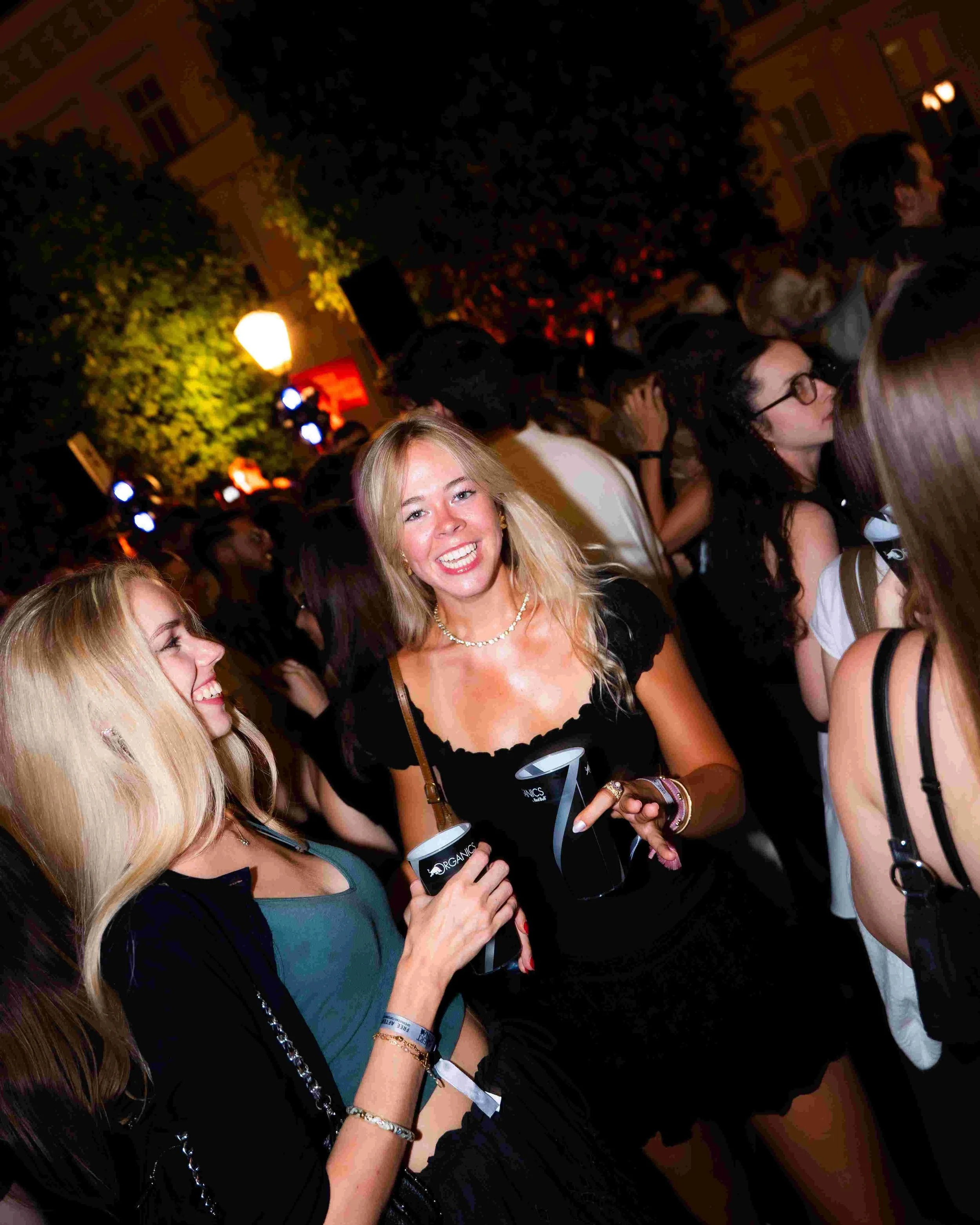 Group of people at an indoor social event, including women holding black cups with 'Organic' printed on them, engaged in conversation, with warm lighting and a lively atmosphere.