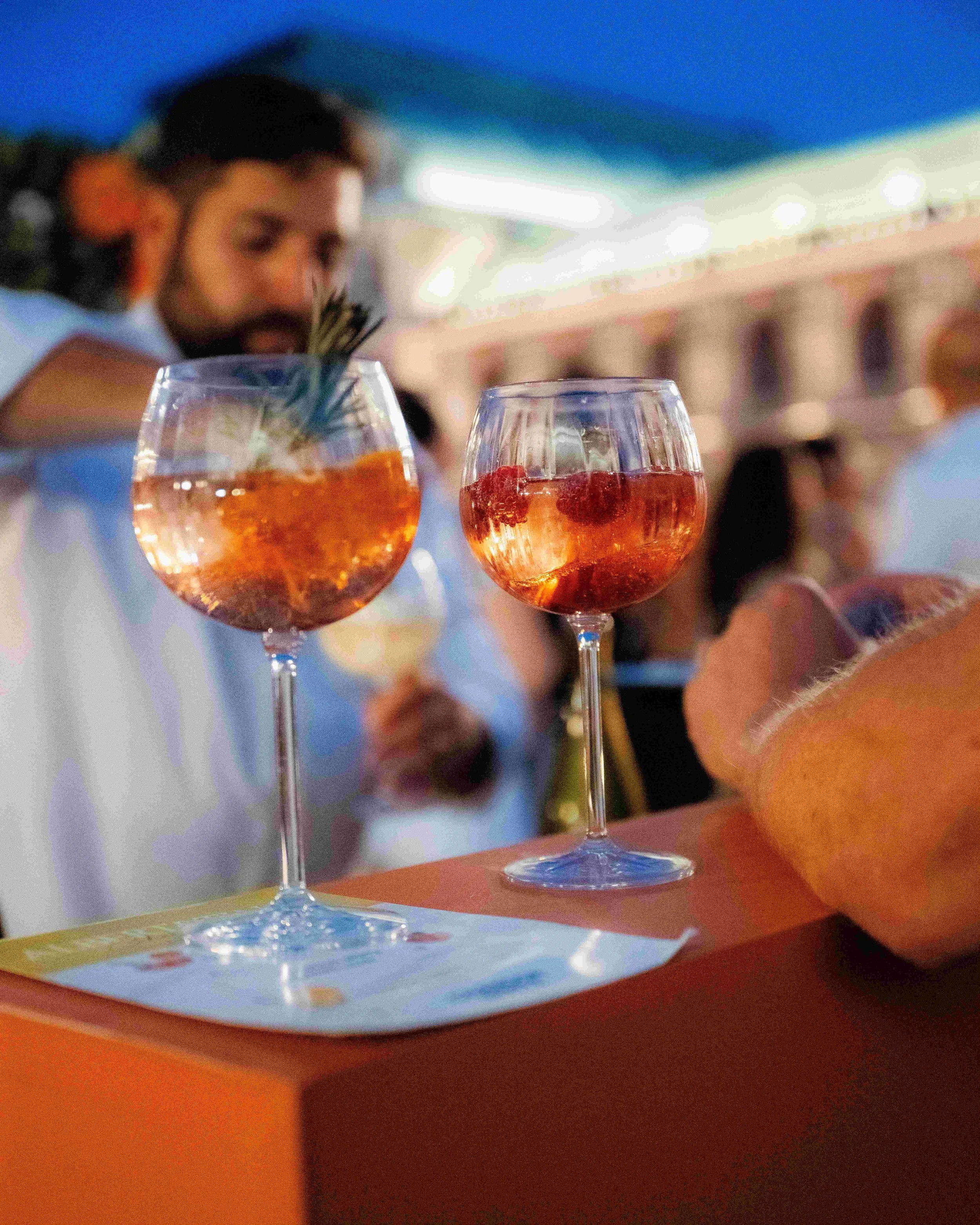 Two glasses of pink-colored beverage with ice and berries, placed on a table. A woman in the background appears to be at a social event or celebration.