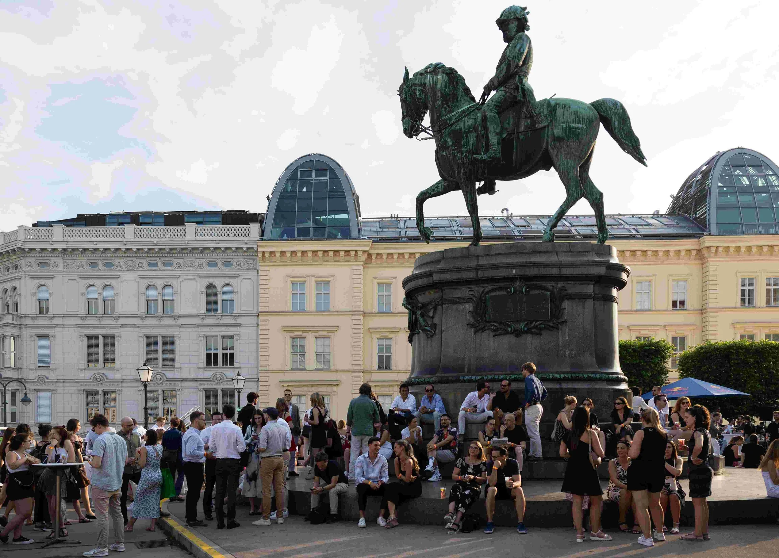 A large group of people gathered in a city square with a bronze equestrian statue of a man in military uniform at the center. Surrounding the crowd are historical buildings, some with modern glass domes, under a cloudy sky.