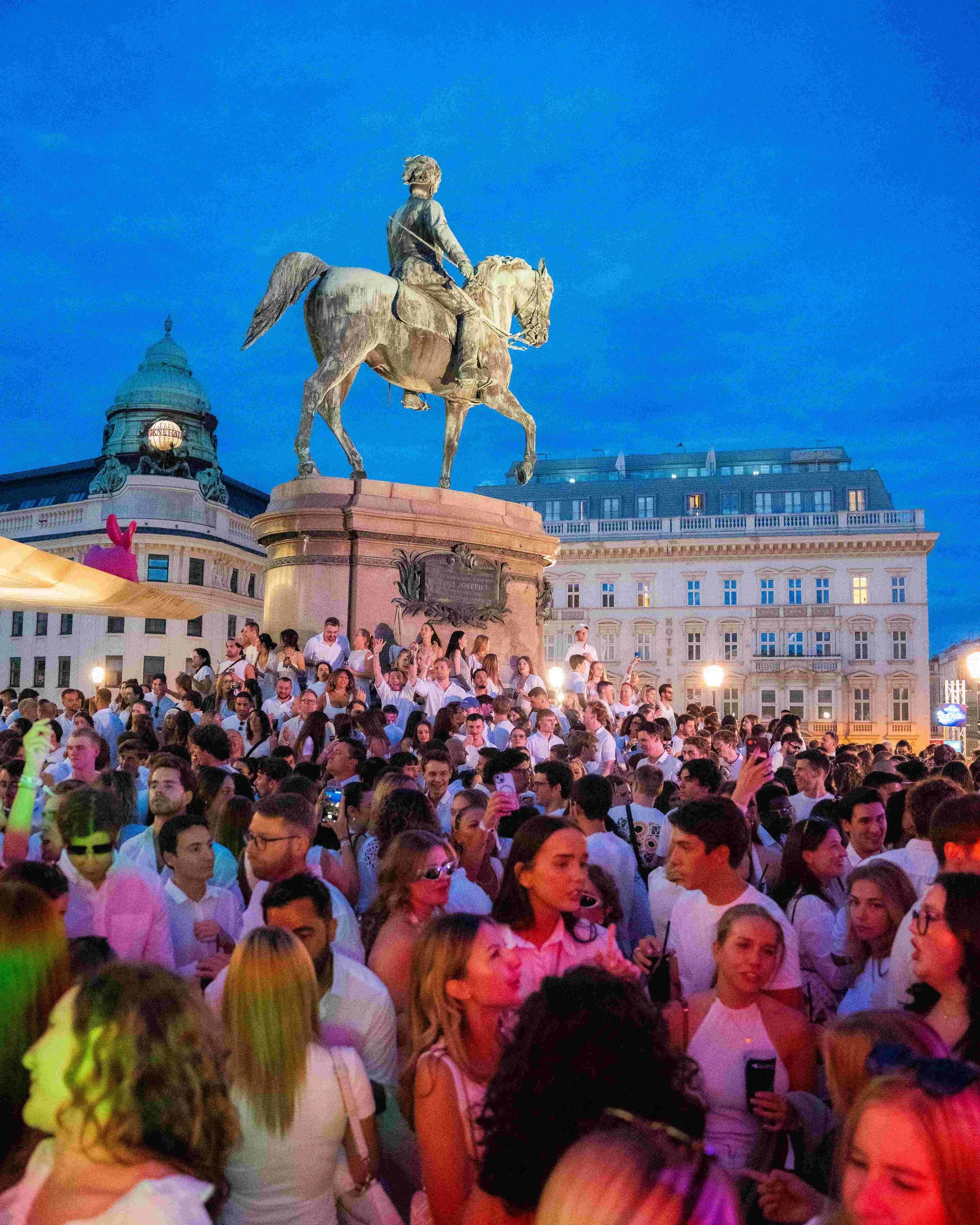 A large crowd of people gathered in front of a historic building with a dome and an equestrian statue on a pedestal, during evening time with a blue sky.