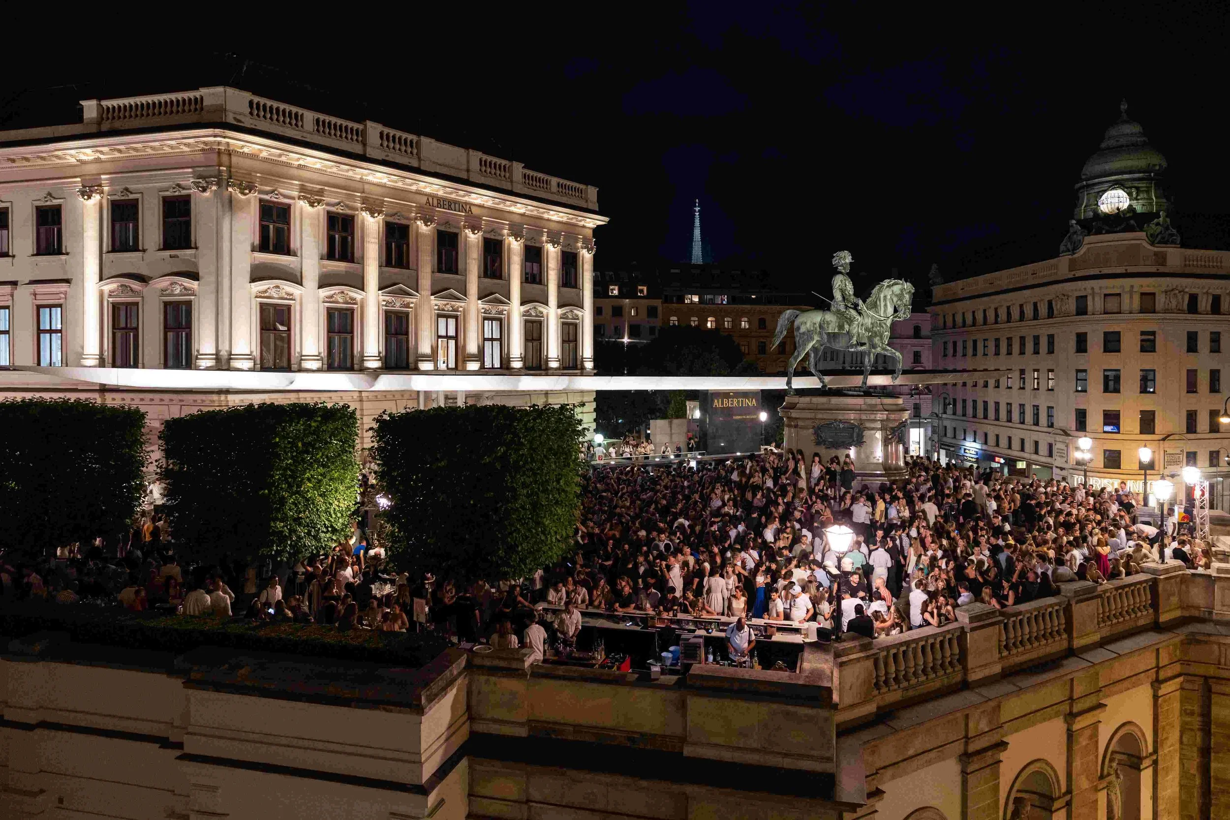 Night scene of a large crowd gathering on an outdoor terrace in front of a decorated building with columns and a statue of a man on horseback, illuminated by lights.