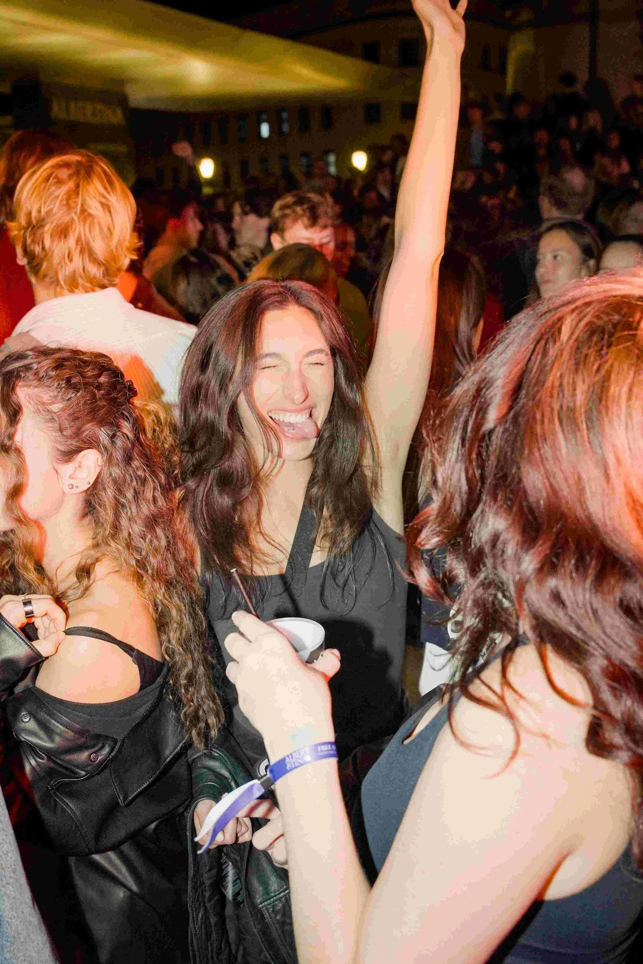 A large crowd of young people at a concert or party indoors. The focus is on a woman with long dark hair who is smiling and raising her hand, surrounded by others with various hair colors and styles.