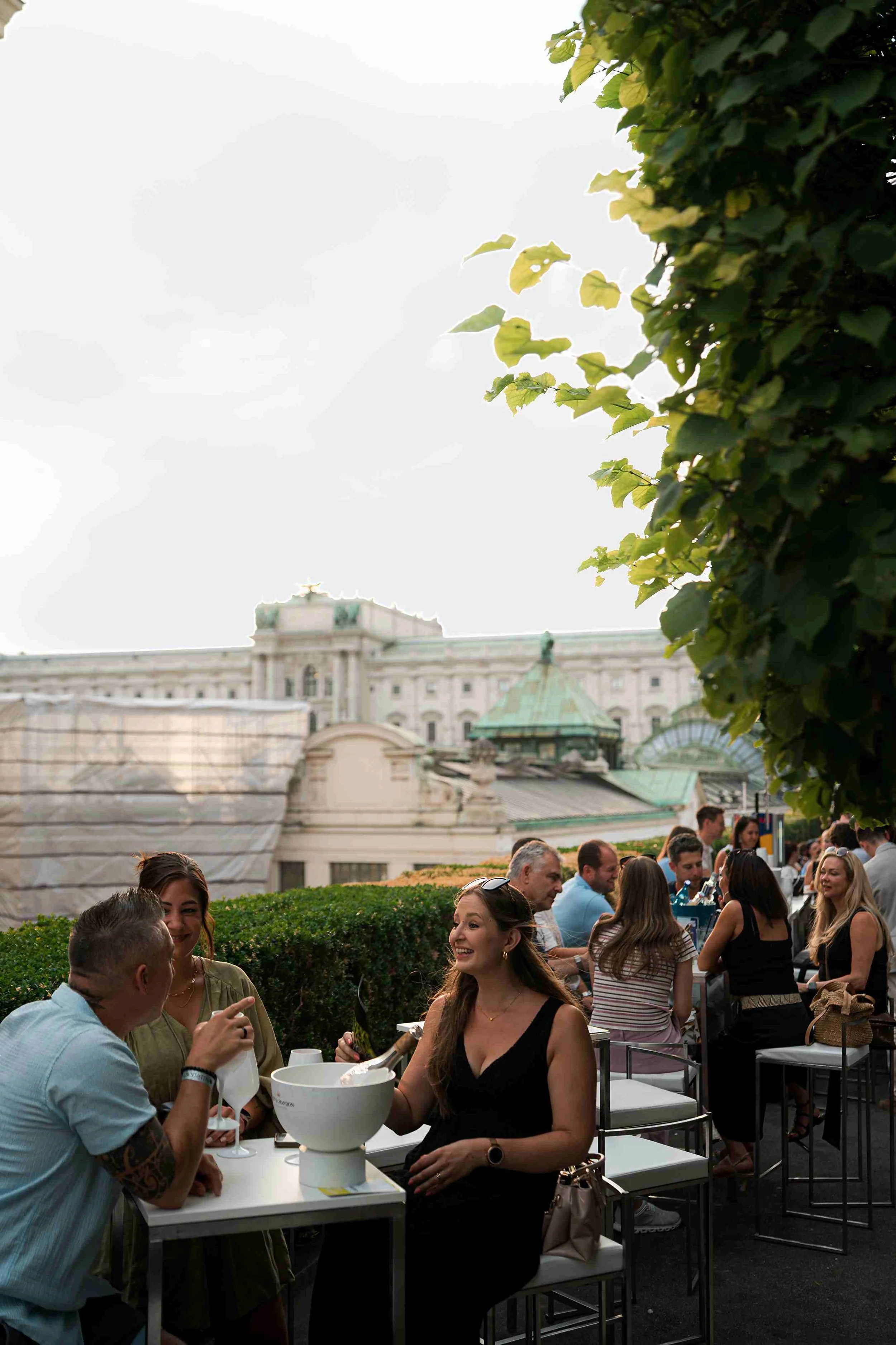 People enjoying a social gathering outdoors at a rooftop venue with a historic building in the background, under a cloudy sky.