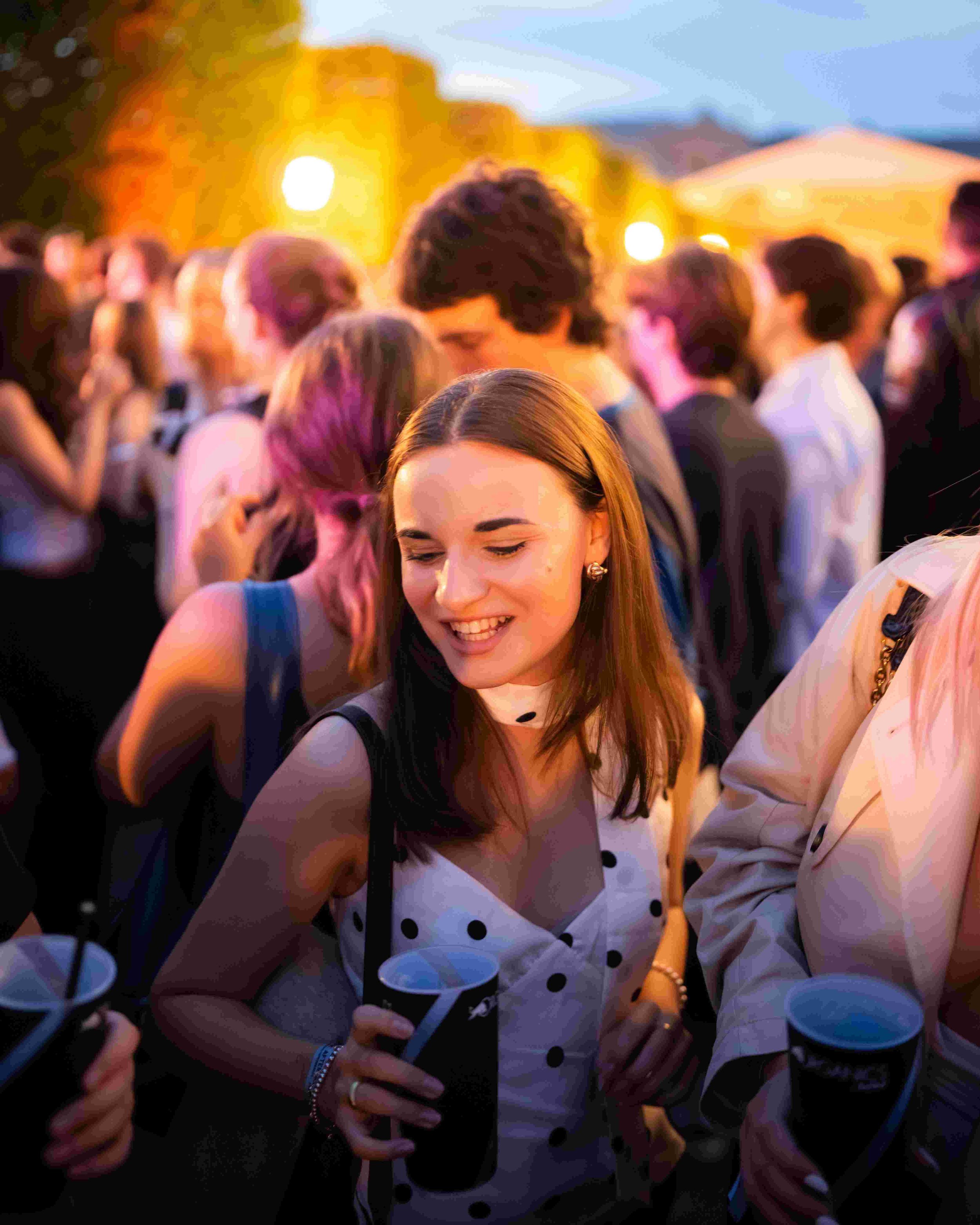 A group of people at an outdoor event during sunset, with a woman in the foreground smiling and looking at her phone, surrounded by others who are socializing and holding drinks.