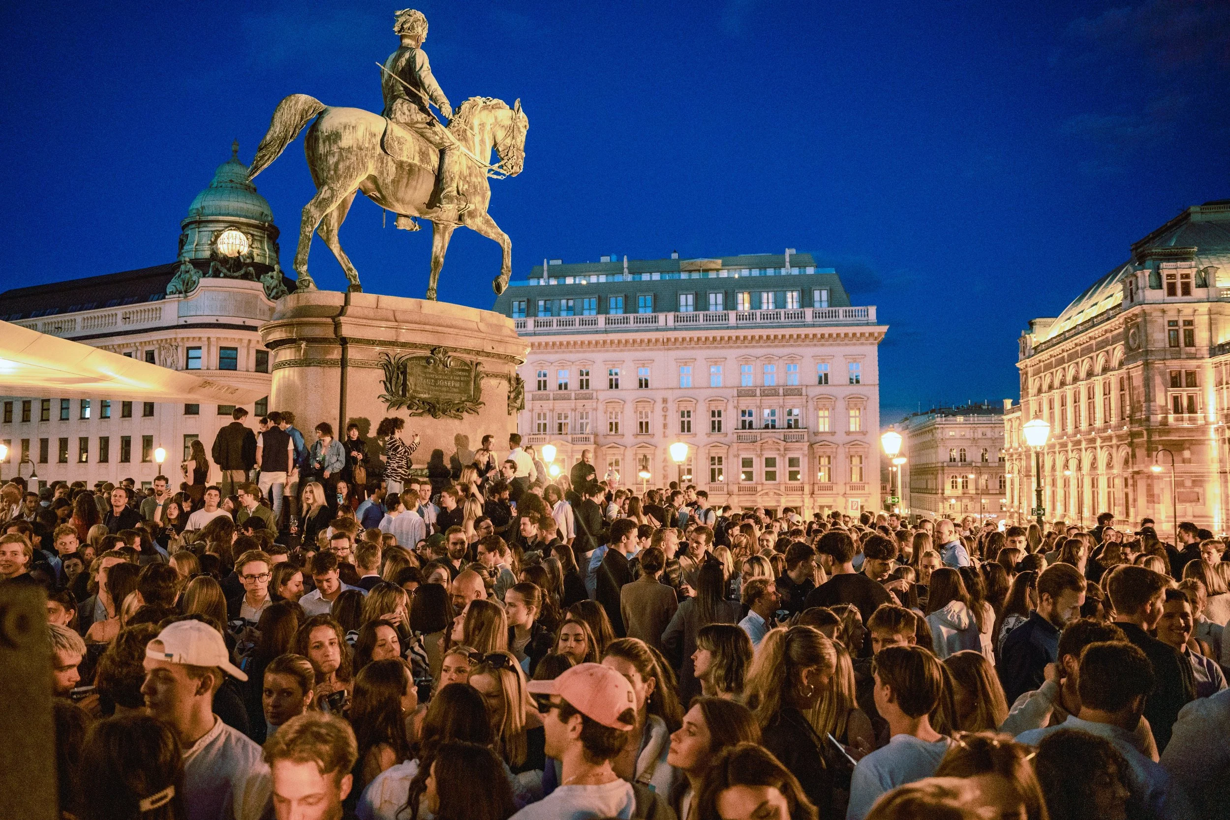 A large crowd of people gathered in an outdoor city square during the evening, with historic buildings and a statue of a man on a horse in the background, illuminated by streetlights.