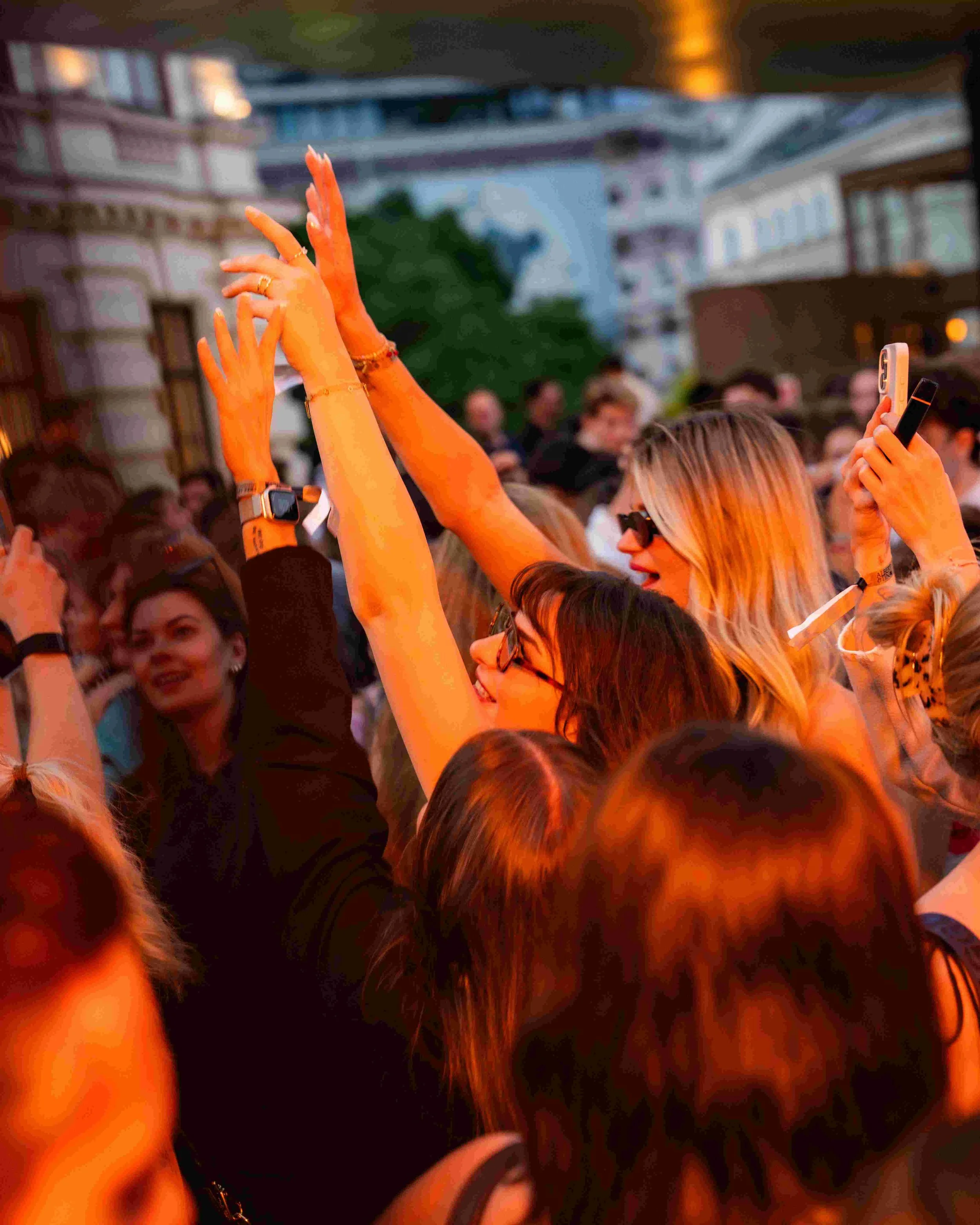 A crowd of people at an outdoor event during sunset, raising their hands and taking photos with their phones.