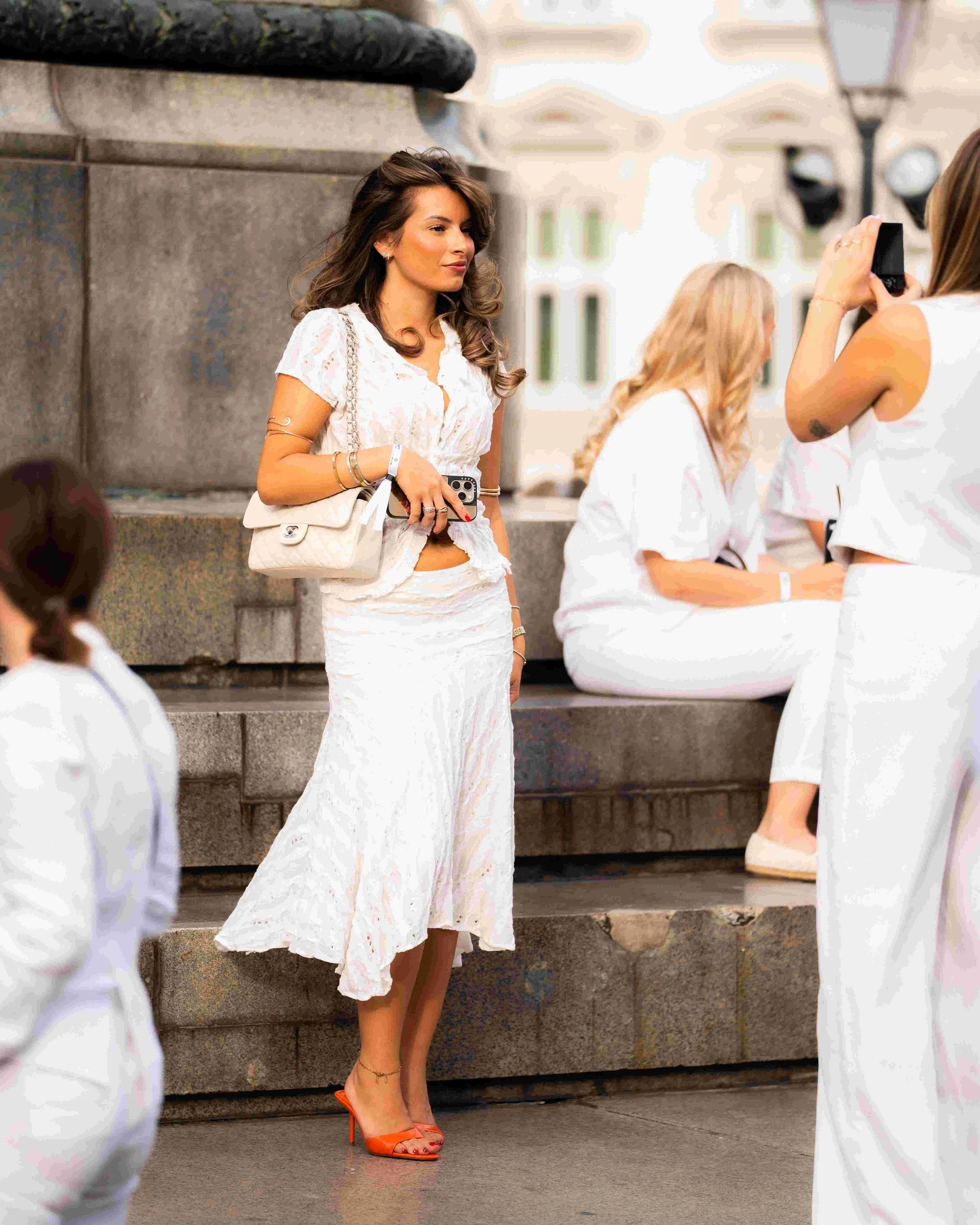 A woman with long, wavy brown hair wearing a white lace dress and orange high heels, standing on stone steps, taking a selfie with another woman sitting on the steps and dressed in white, in an outdoor urban setting.