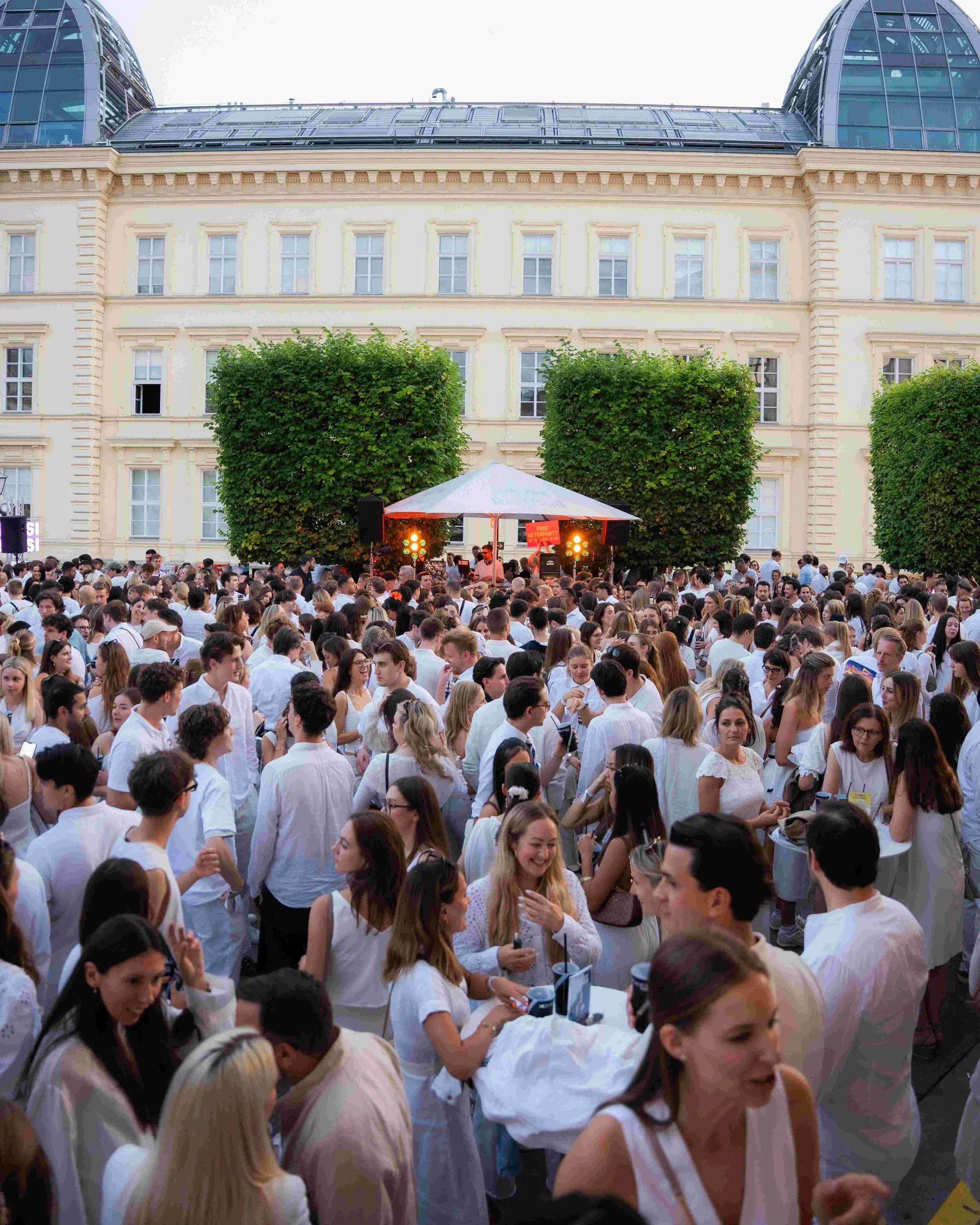 Crowd of people attending an outdoor event, some wearing white clothing, with a stage in the background and large green trees flanking the stage. The building behind has multiple windows and a glass dome roof.