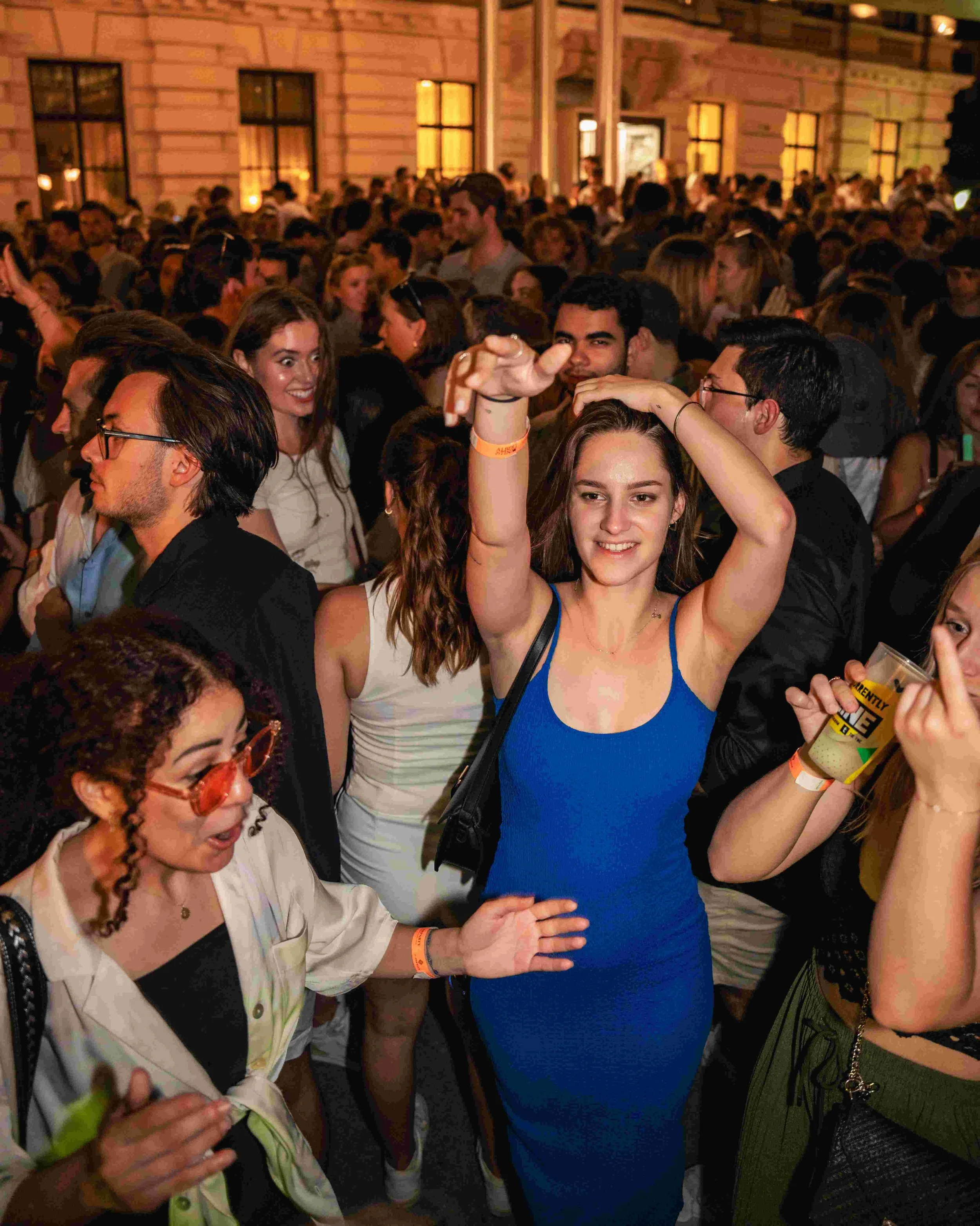 People socializing at an indoor event, with a woman in a blue dress in the foreground holding her arm up and smiling.