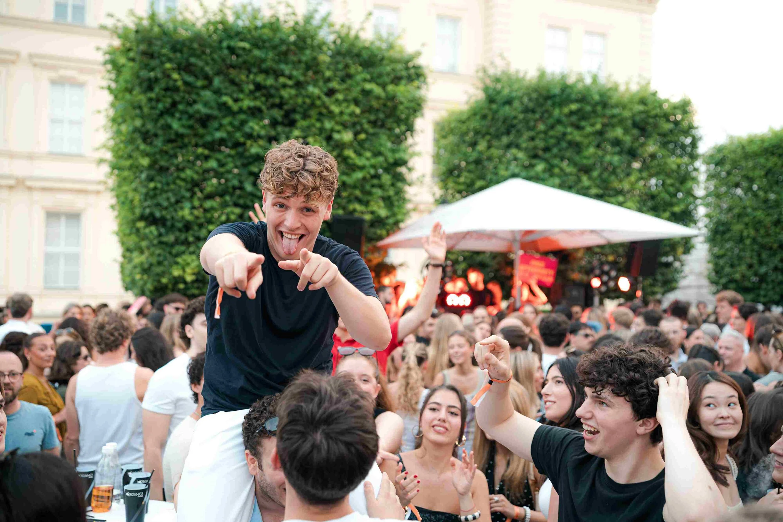 A young man with curly hair is sitting on someone's shoulders, smiling and pointing towards the camera at an outdoor gathering or concert. The crowd is dense with many people smiling and enjoying the event, and there is a stage with lighting and a la