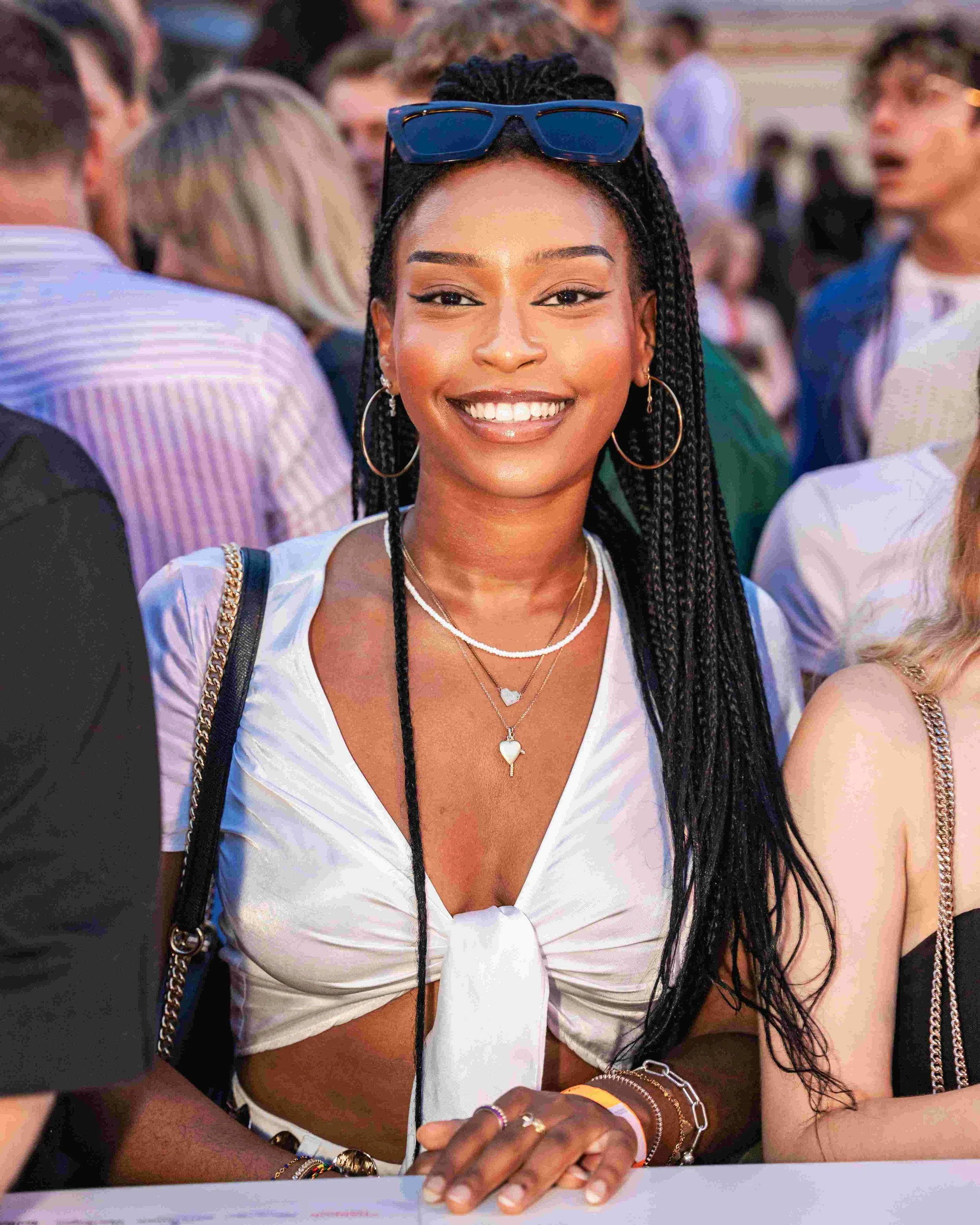 A young African American woman with long braided hair, wearing sunglasses on her head, smiling warmly, dressed in a white crop top, in a crowded social setting.