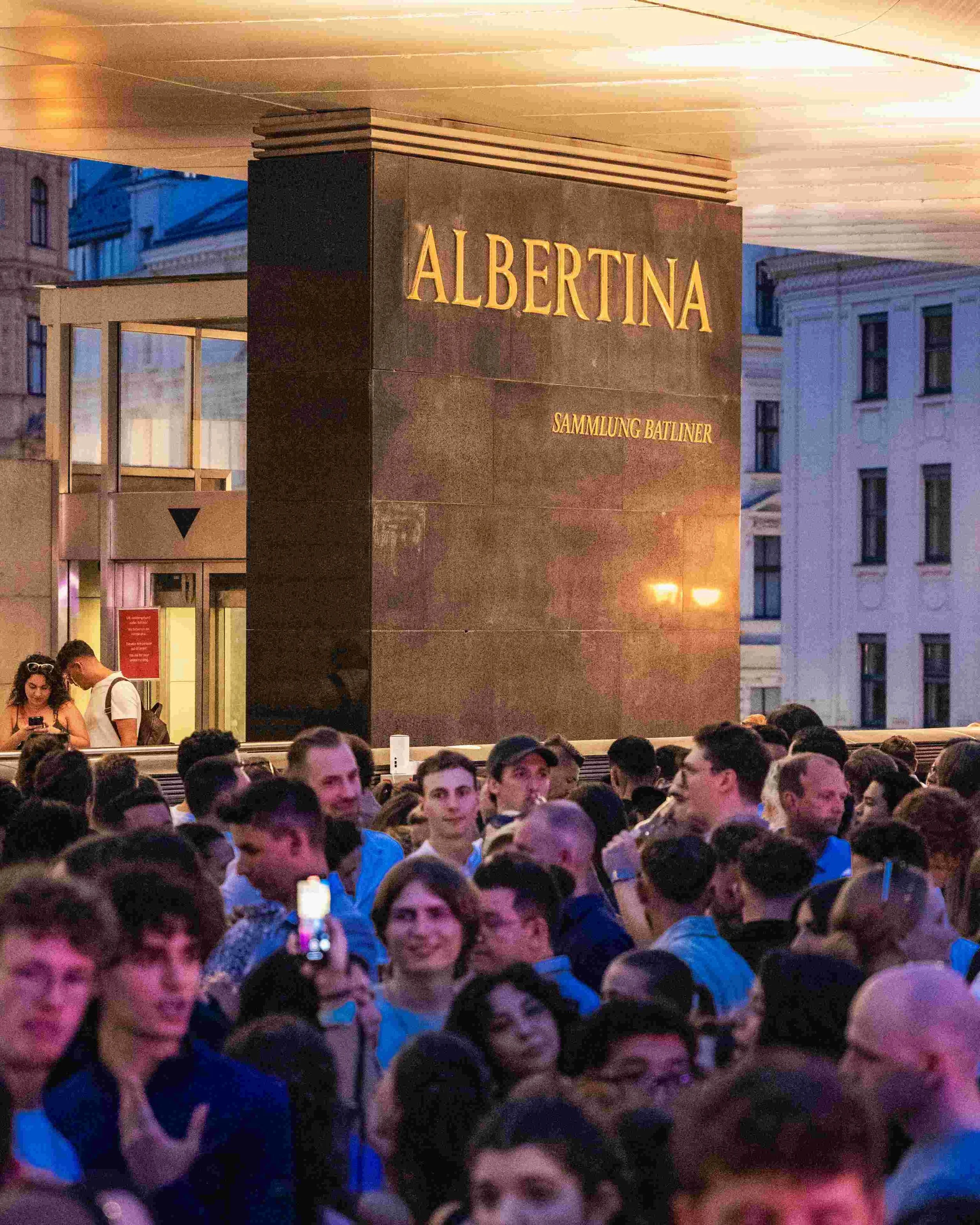 A large crowd of people gathered outside a building with a sign that reads 'Albertina Sammlung Batliner' during the evening. The crowd includes young adults and the scene is set in an urban area with other buildings visible in the background.