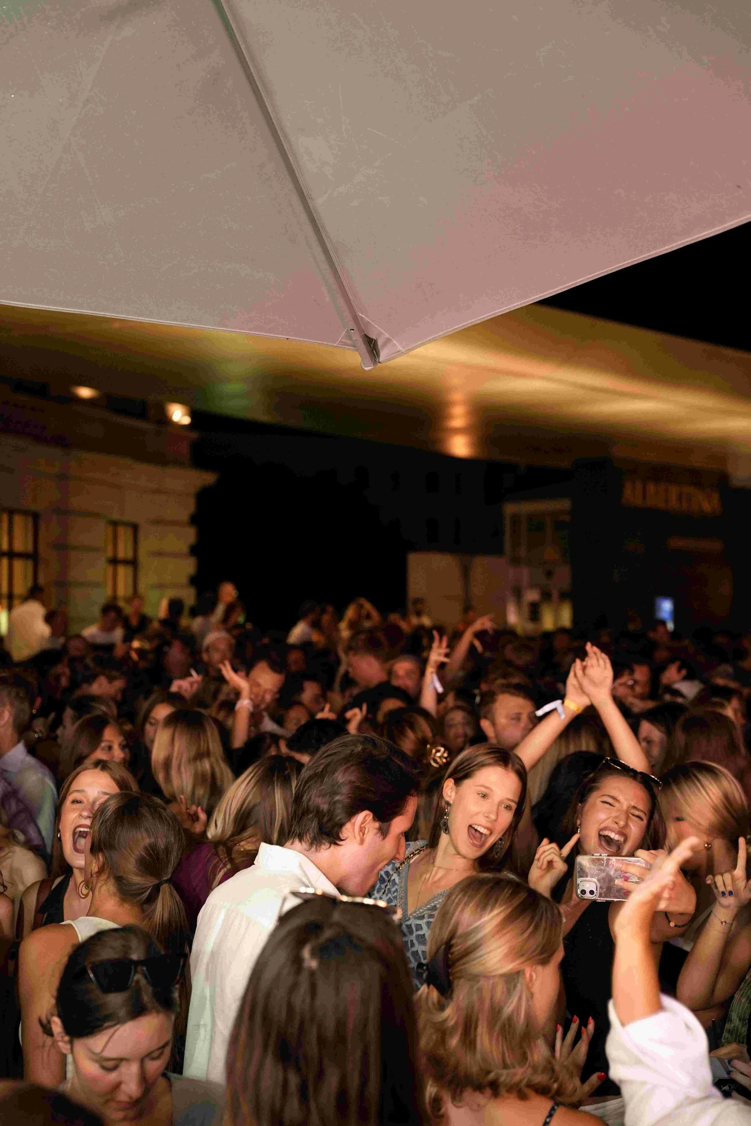 Crowd of people dancing and celebrating at an indoor event, many smiling and raising their hands, with a blurred background of a decorated room.