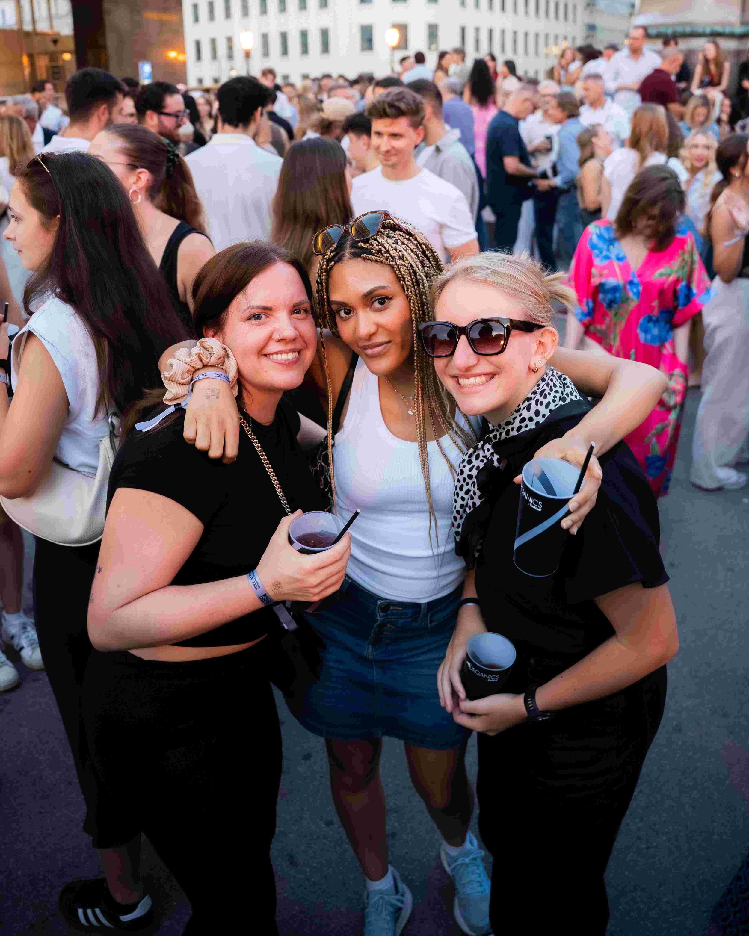 Three women are smiling and hugging in a crowded outdoor event or party at dusk with many people in the background. They are holding drinks and appear happy.