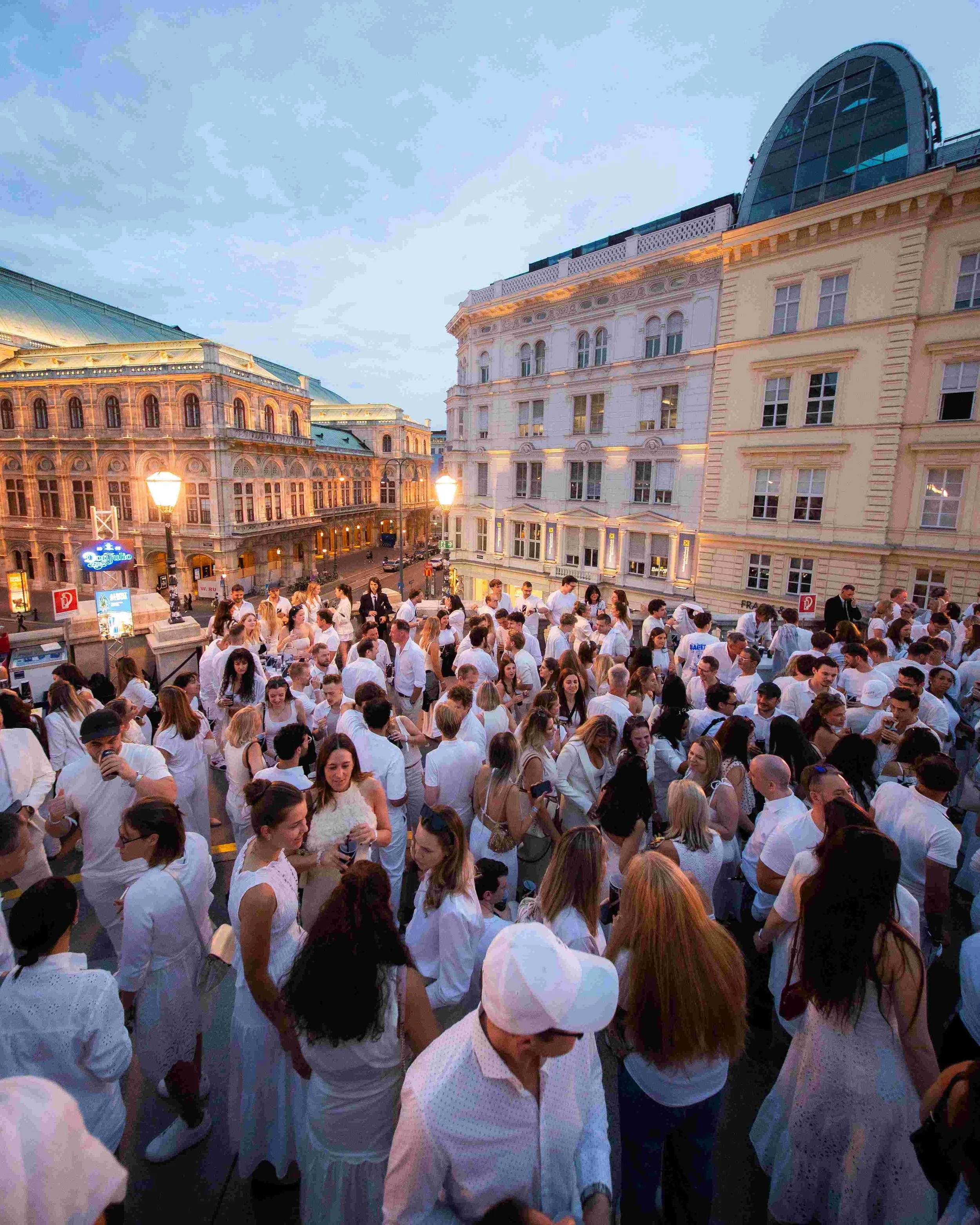 Crowd of people gathered outdoors near historic buildings during early evening, with some people wearing white shirts and glasses, and street lamps illuminating the area.