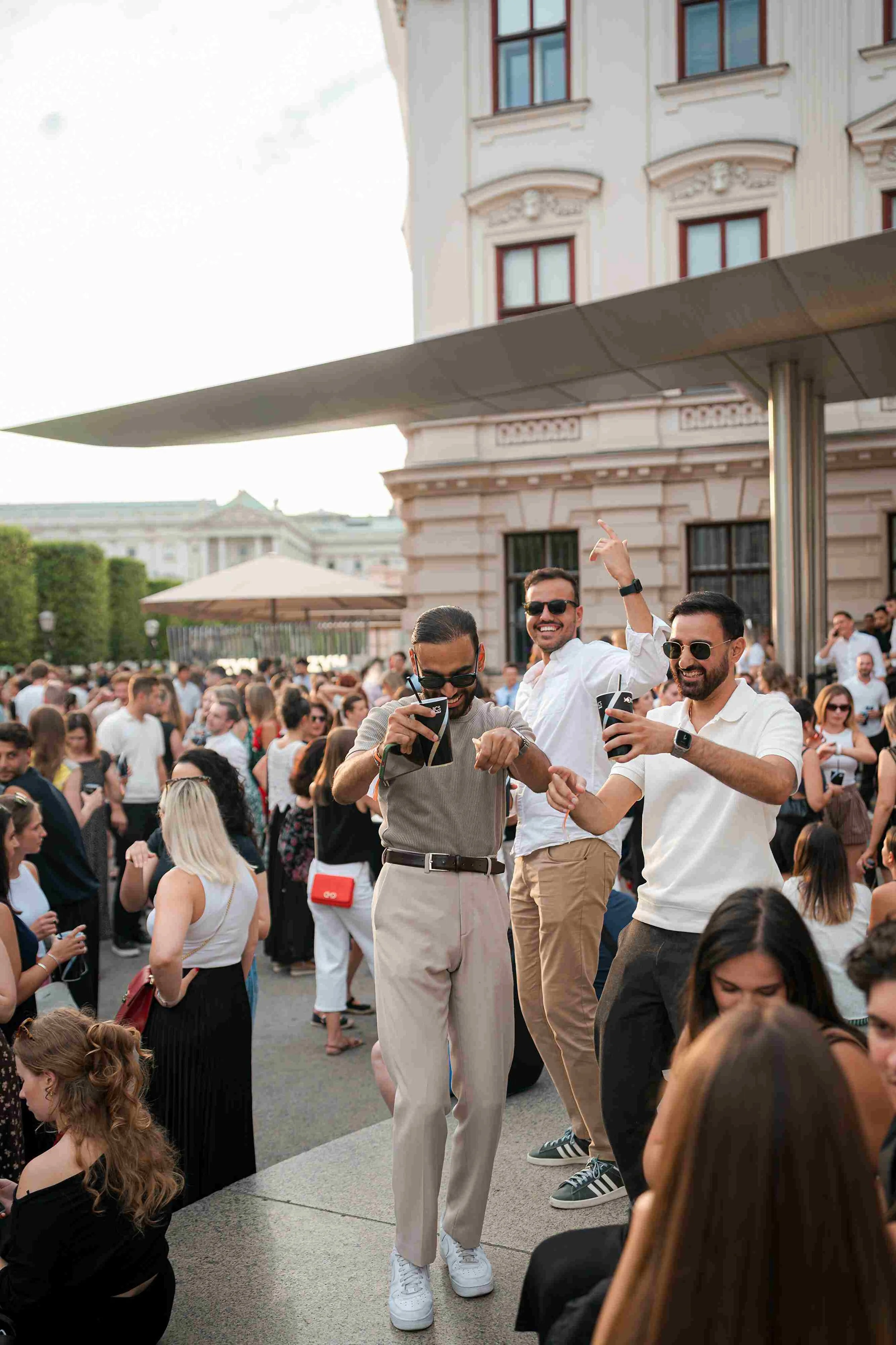 A group of four men enjoying at a large outdoor event. They are standing on a stage with a crowd behind them. The men are dressed in casual attire, with three wearing sunglasses. One of the men is wearing a white shirt, and others are wearing beige a