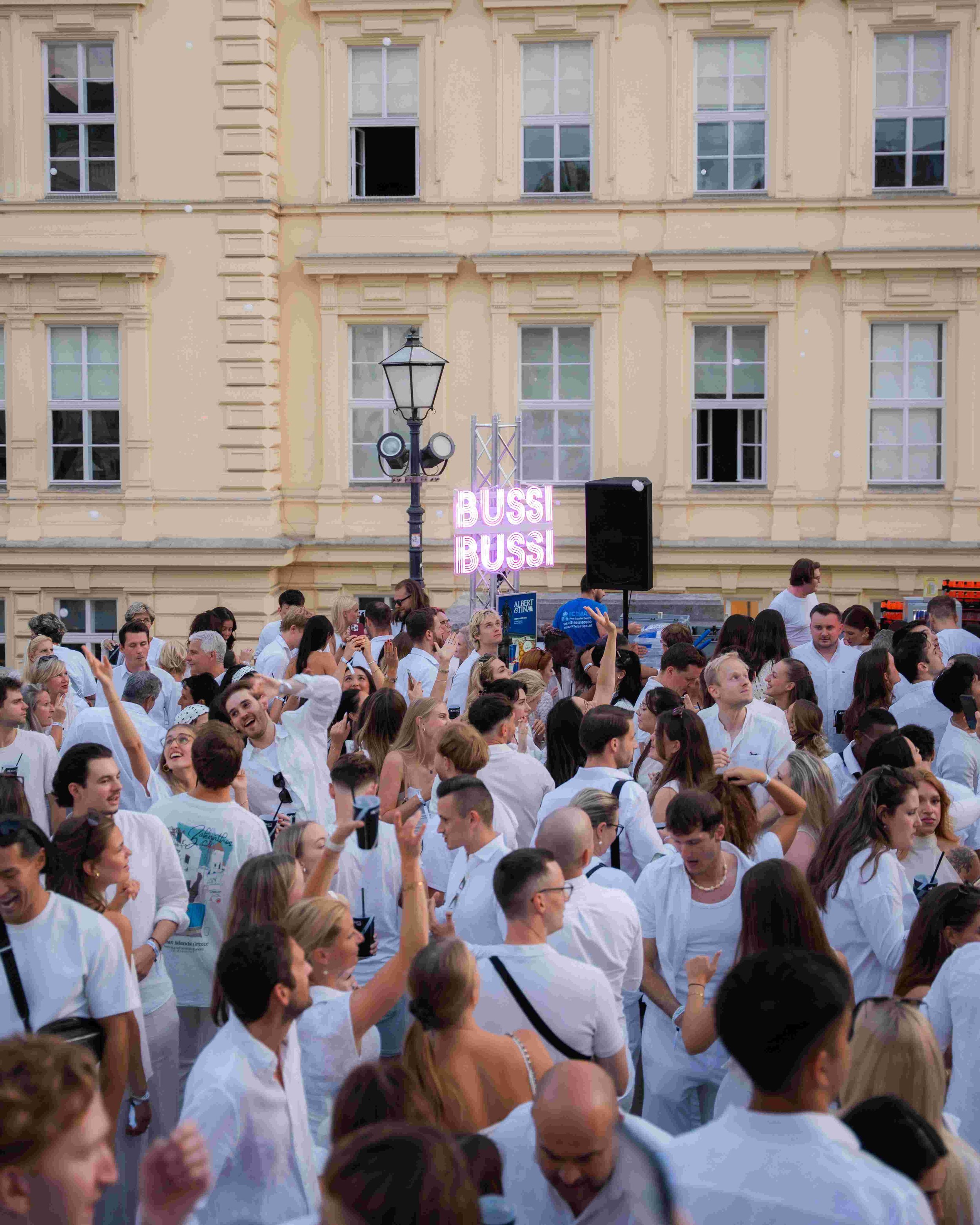 Crowd of people dressed mostly in white gathered outdoors in front of a multi-story beige building with multiple windows. There is a pink neon sign with the word 'BUSS' visible in the background, along with some audio equipment and a street lamp.