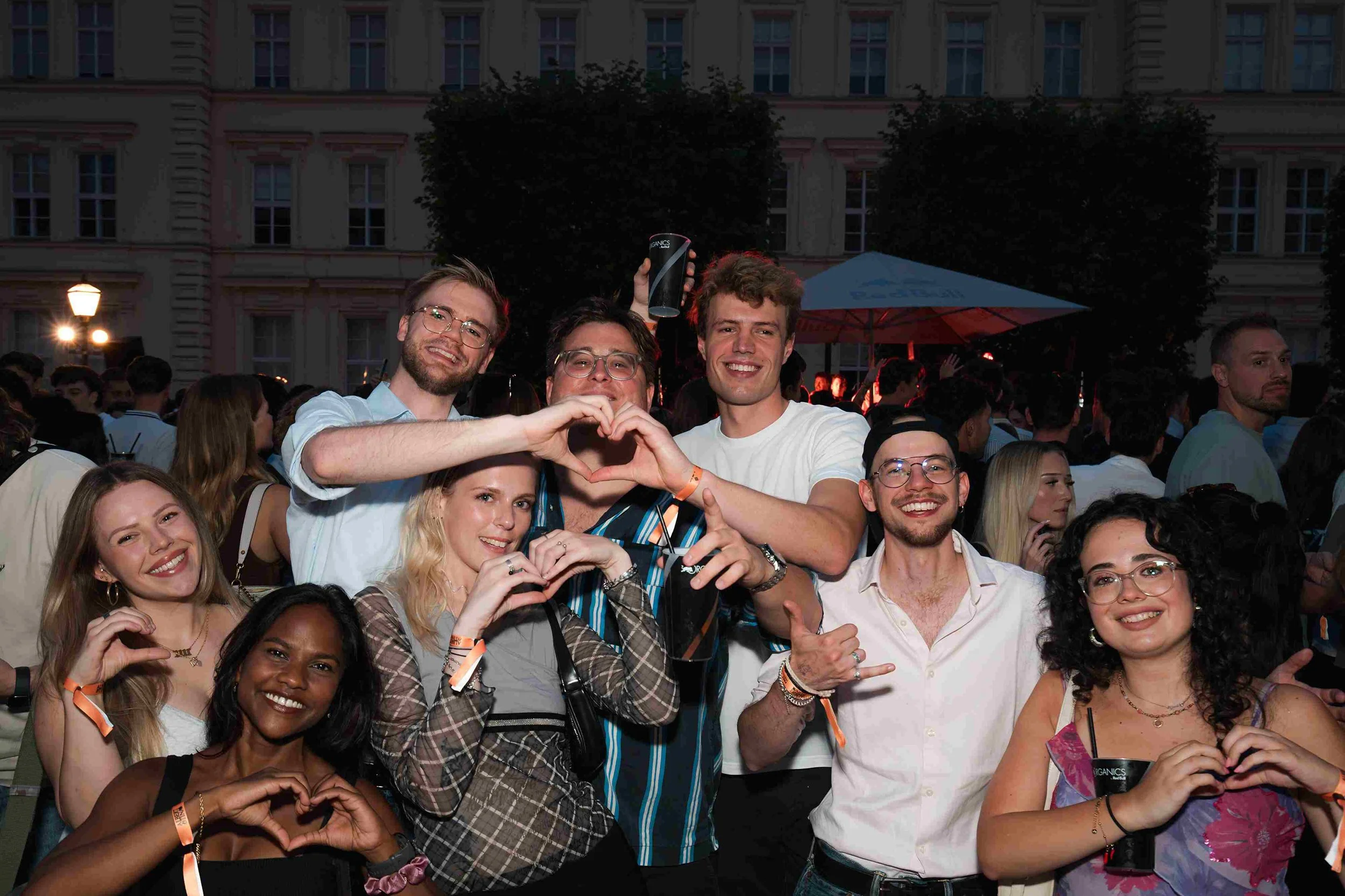 A group of people at an outdoor event during the evening, smiling and making heart shapes with their hands, with some wearing orange wristbands.