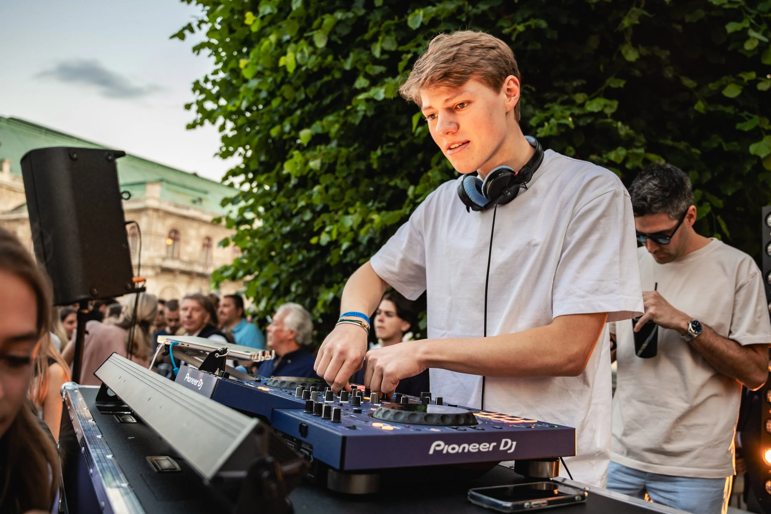 A young man with headphones around his neck DJing at an outdoor event with a crowd of people gathered around.