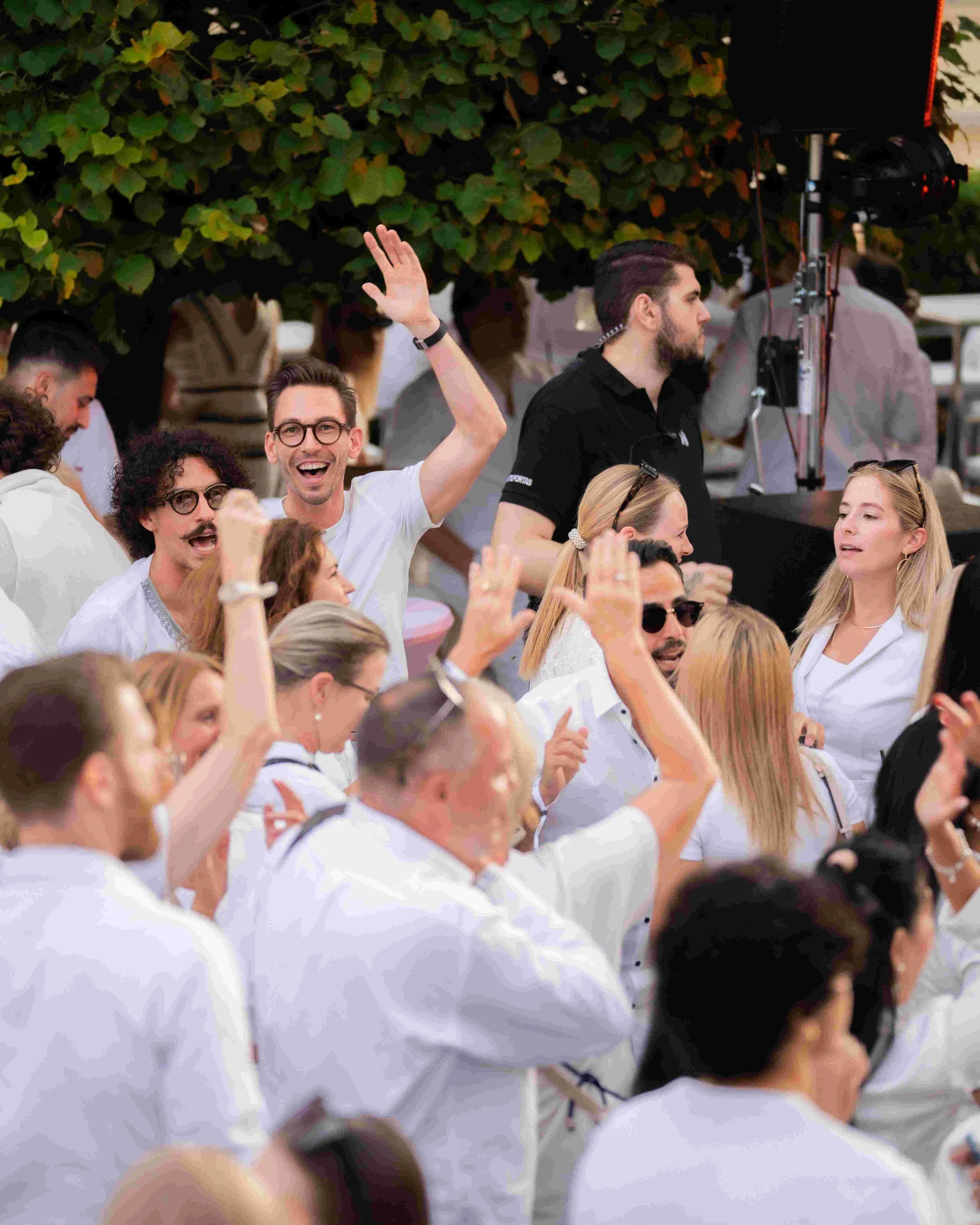 Group of people gathered outdoors, some smiling and engaging with each other, with lush green trees in the background.