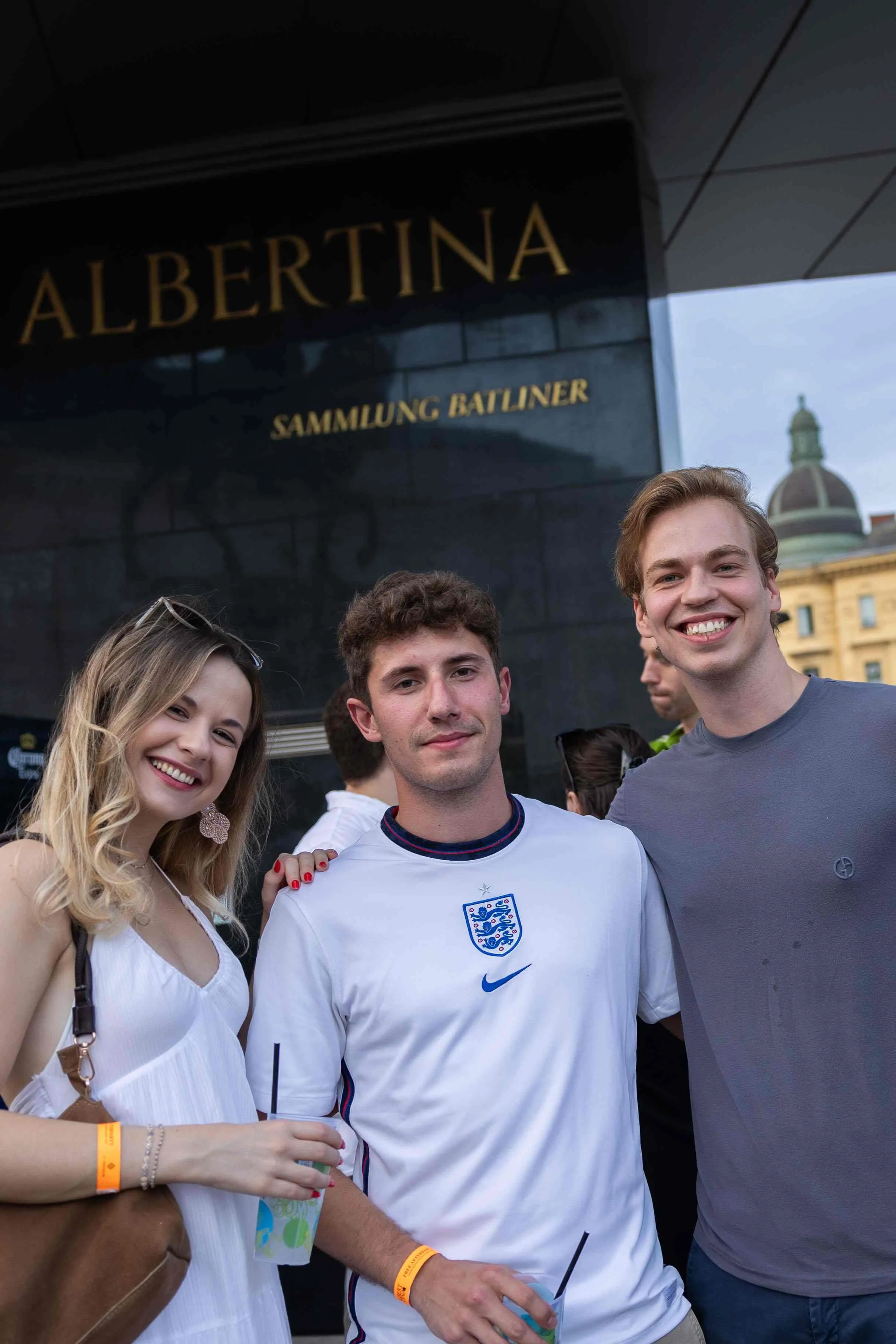 Group of young adults smiling and posing for a photo in front of a building with the sign 'Albertina Sammlung Batliner' and a church in the background. One person is wearing a white England football jersey.